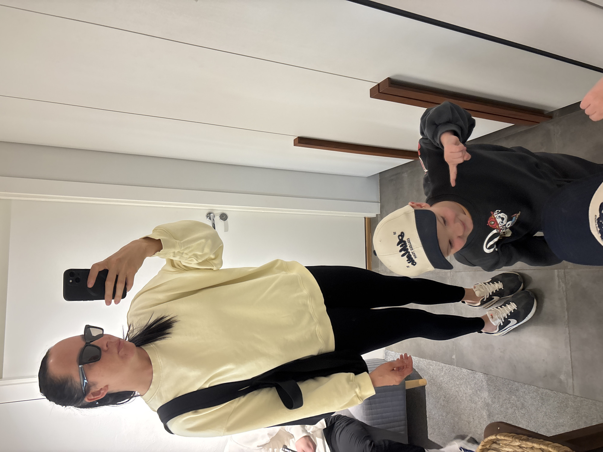 A woman in sunglasses and a yellow sweatshirt and black leggings is taking a mirror selfie with her black cell phone. She has a black strap across her torso. Next to her, a child in a black sweatshirt and a baseball cap is giving a thumbs down. They are standing in a room with white walls and grey tile floor. There is a wooden bar on the wall behind them. The location is not identifiable as Chaikovsky, Russia.