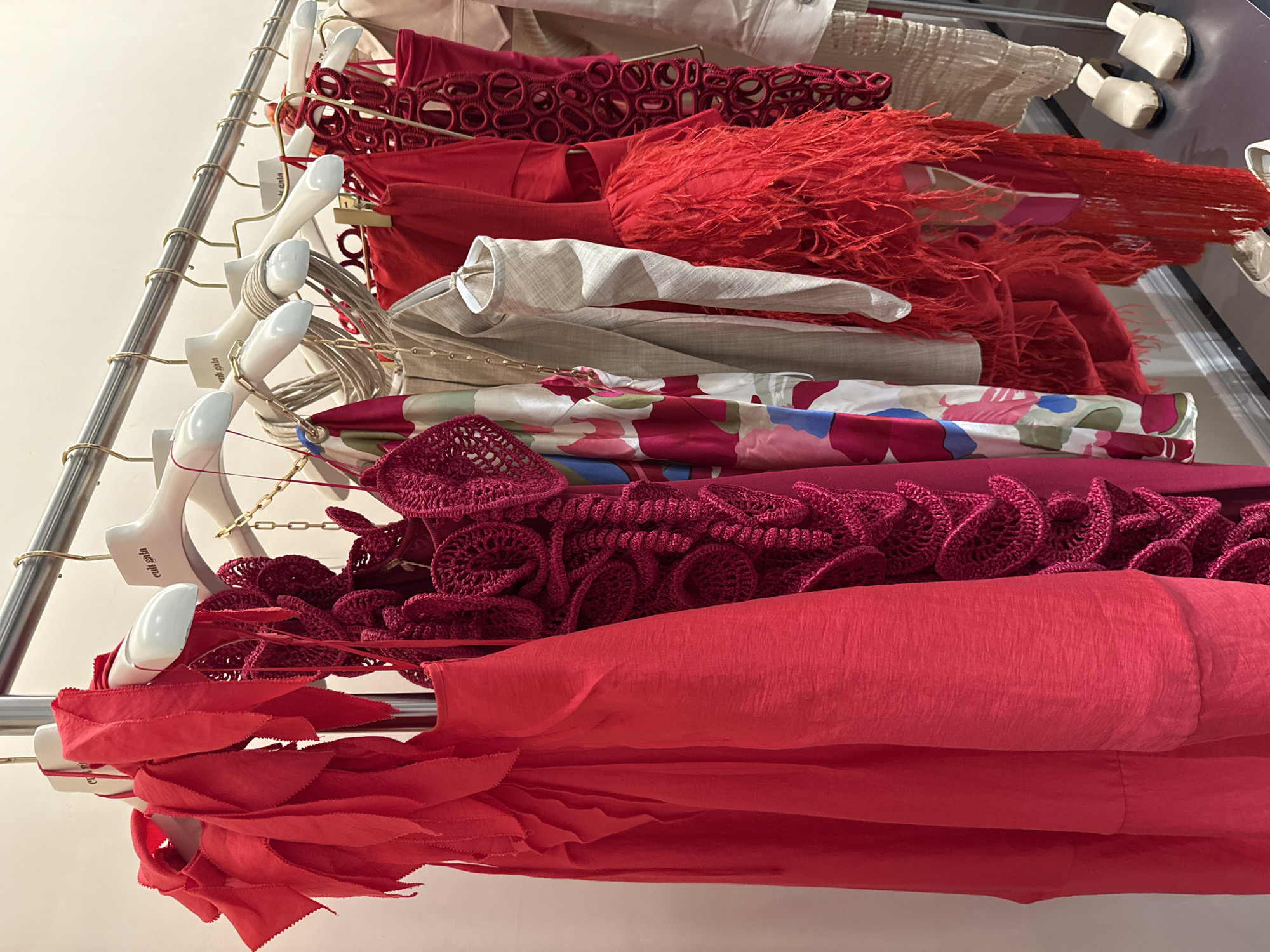 The image shows a clothing rack with various garments in shades of red, beige, and floral prints, against a light-colored wall. The garments are hanging on white hangers, and the rack itself is made of metal. From top to bottom, the garments appear to be: a beige garment, a red garment with circular cutouts, a red garment with fringe, a beige jacket, a silk garment with floral designs and red, crochet garments and a red garment with layered frills. The location appears to be inside a clothing store or studio in Sunrise, United States of America. There are no people visible.