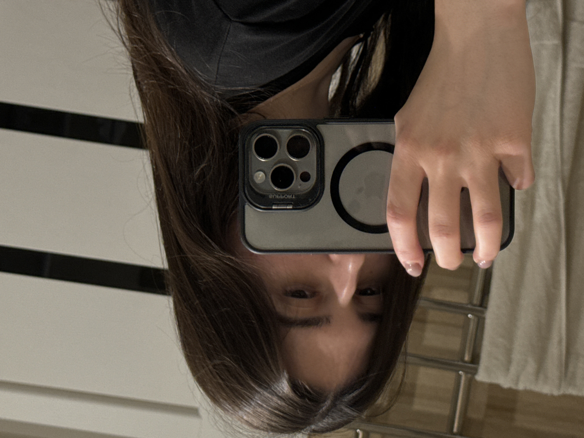 This is a selfie taken by a young woman with long brown hair. She is wearing a black t-shirt and holding a smartphone in her left hand, which is angled to capture her reflection in a mirror. The phone has a triple-lens camera and a circular magnetic attachment on the back. The background appears to be a bathroom, with a towel rack and a horizontally striped wall visible. The lighting is dim. There are no discernible landmarks or elements that would specifically place this image in Vinnytsia, Ukraine.