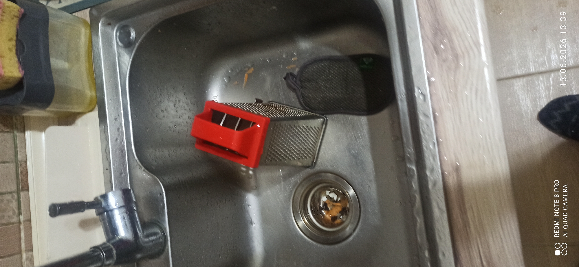 The image shows a close-up view of a stainless steel sink. In the sink, there is a red and silver box grater, and a dark grey mesh scrubber. The sink drain is visible and contains some food debris. A faucet is partially visible on the left side of the sink, and a soap dispenser with a sponge is to the right, behind the sink. The surrounding countertop appears to be made of wood-patterned laminate. The image is a photograph taken from a slightly elevated angle, capturing a domestic kitchen scene.