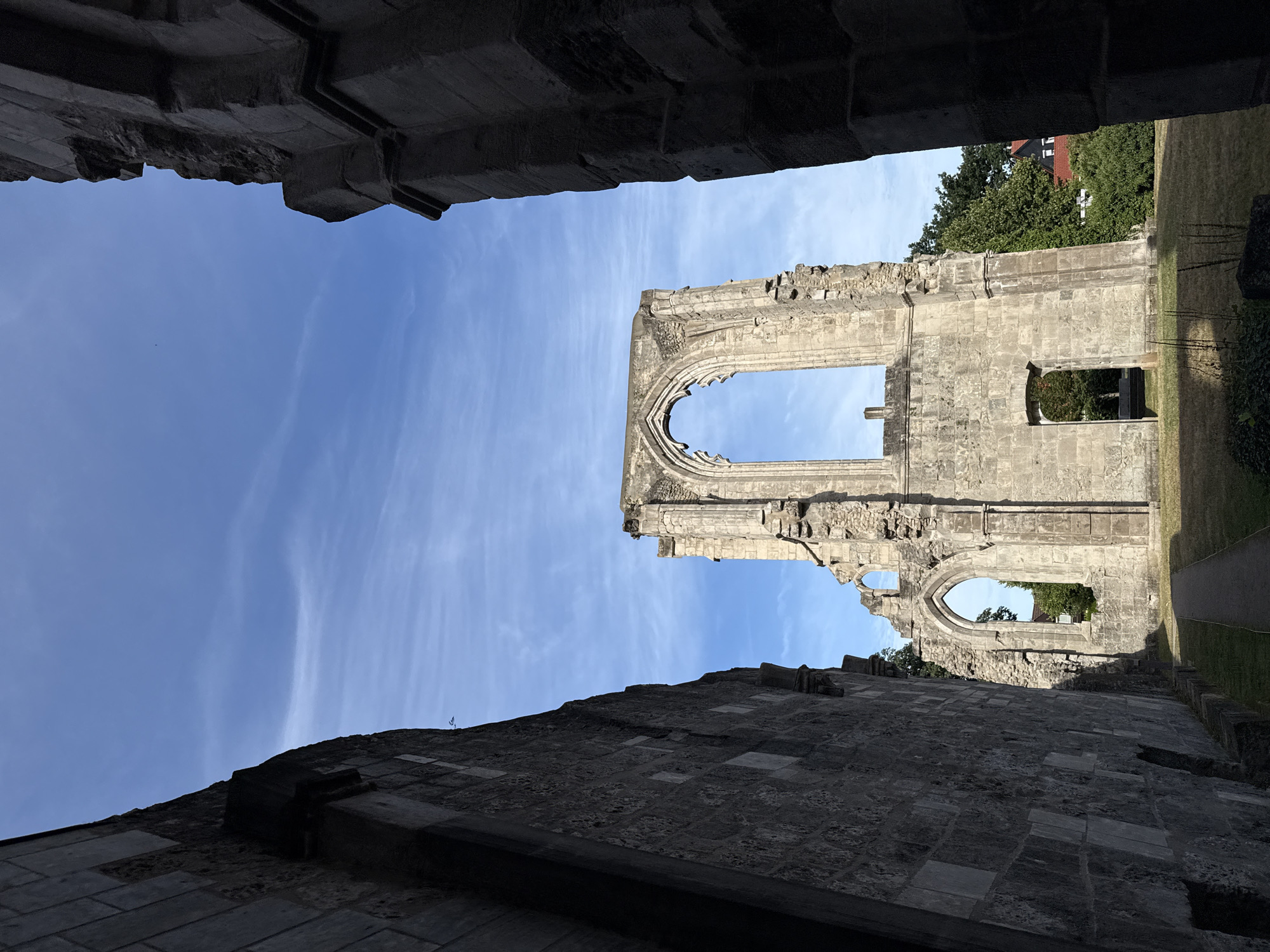The image shows a view of the ruins of the Walkenried Abbey in Walkenried, Germany, on a clear day with a blue sky and scattered white clouds. The foreground is dominated by the dark, weathered stone walls of the abbey ruins, framing a central view of a large, arched window opening in a towering section of the structure. The stone is rough and textured, showing signs of age and decay. Sunlight casts shadows across the scene, highlighting the architectural details of the crumbling stonework. Beyond the main ruin, green grass and trees are visible, with glimpses of a red-tiled roof and dark buildings suggesting the proximity of a town. A paved pathway leads into the ruins, and the overall impression is one of historical grandeur and natural decay.