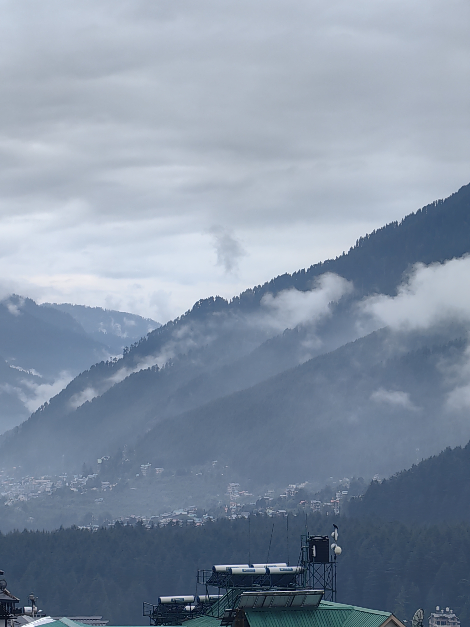 This is a vertical, full shot image of the mountainous landscape surrounding Vashisht, India, under a cloudy, overcast sky. The scene is dominated by rolling hills covered in dense pine forests, with several layers of these hills receding into the distance. Wisps of fog and mist cling to the slopes, partially obscuring the trees and creating a soft, hazy effect. A small town or village is visible nestled on the side of one of the hills, with numerous buildings clustered together. In the foreground, the rooftops of several buildings are visible, some equipped with solar water heaters and antennas. One building has a prominent water tank and communication equipment on its roof. The overall impression is one of a serene, mountainous environment with signs of human habitation.
