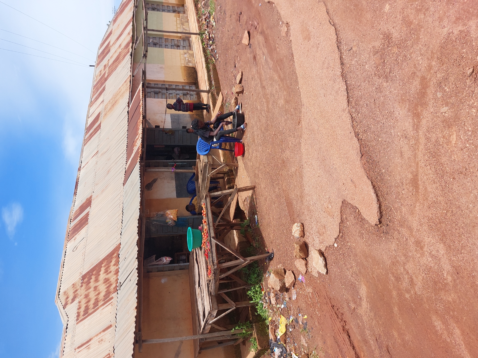 The image depicts a scene in Barakin, Nigeria, featuring a weathered, corrugated metal-roofed building with a brown and ochre facade. In front of the building, a wooden stall displays red fruits, possibly tomatoes, and a large green basin. A young girl, wearing dark clothing, sits on a blue plastic chair to the right of the stall, attending to a black bowl. Another child, dressed in a red and dark patterned garment, stands further back, observing the surroundings. The ground in the foreground is a mix of reddish-brown dirt and gravel, with scattered rocks and debris. A portion of a paved road is visible in the lower right of the frame. The sky above is a clear blue with a few wispy clouds. The overall impression is one of a humble market or roadside stall in a rural or semi-urban setting.