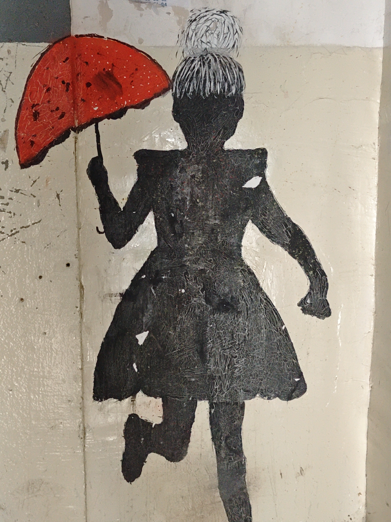 A mural depicting a silhouette of a girl in a dress holding a red umbrella against a beige wall. The girl is rendered in dark grey with a textured appearance, and her hair is styled in a bun on top of her head with white accents. The umbrella is red with white polka dots. The wall has a slightly rough texture with some scuff marks and peeling paint. The girl appears to be in motion, with one leg bent as if running.