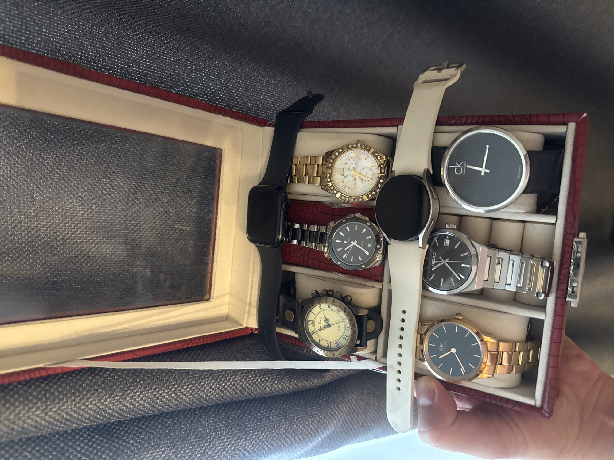 A top-down shot shows a collection of watches inside a red watch box. The watch box is positioned on a textured, gray fabric. The watch box has a mirrored lid which reflects the interior of the box. Inside the box, there are seven watches.
On the top row, from left to right, is a vintage-style watch with a cream-colored dial and Roman numerals. Next to it is a black smartwatch with a black band. To the right of the smartwatch is a gold-tone watch with a white dial, Roman numerals, and diamond markers.
On the bottom row, from left to right, is a gold-tone watch with a dark blue dial and rose gold indices, a silver-tone watch with a black dial and silver indices, and a black watch with a minimalist black dial featuring the Calvin Klein logo and white hands.
A hand is holding the watch box from the bottom, with its thumb visible on the left side. The background consists of a gray textured fabric, possibly a curtain or clothing.
The scene does not provide any information that could be used to determine its location in Vladivostok, Russia.