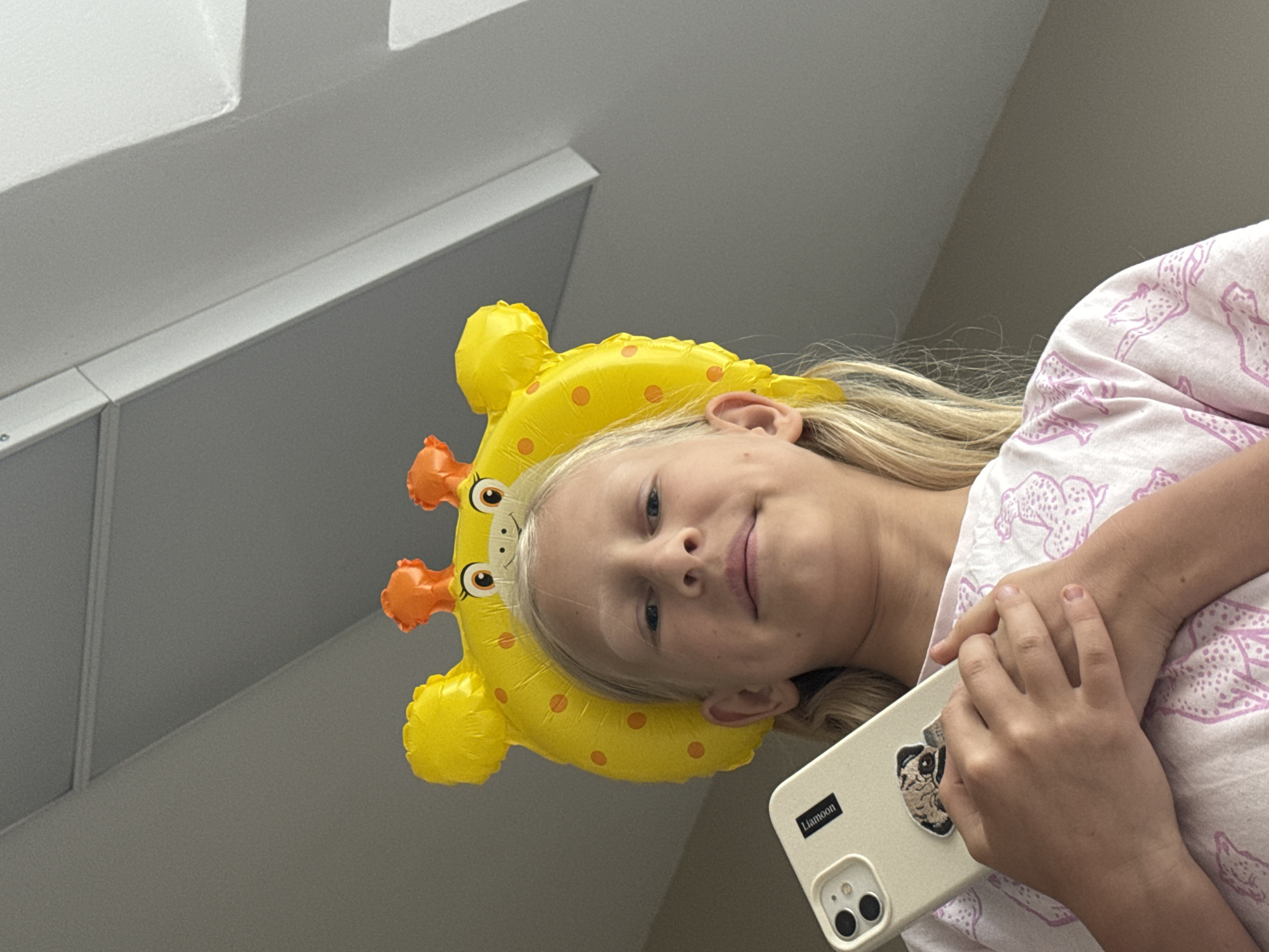 A fair-skinned young girl with blonde hair is oriented at an angle in the frame. She is wearing a yellow giraffe-shaped balloon on her head. She holds a cream-colored smartphone, adorned with a black rectangular label reading "Liamoon," with her hands resting on a pink and white patterned garment. The background consists of a light gray ceiling with white architectural elements. The location or city is indeterminable.