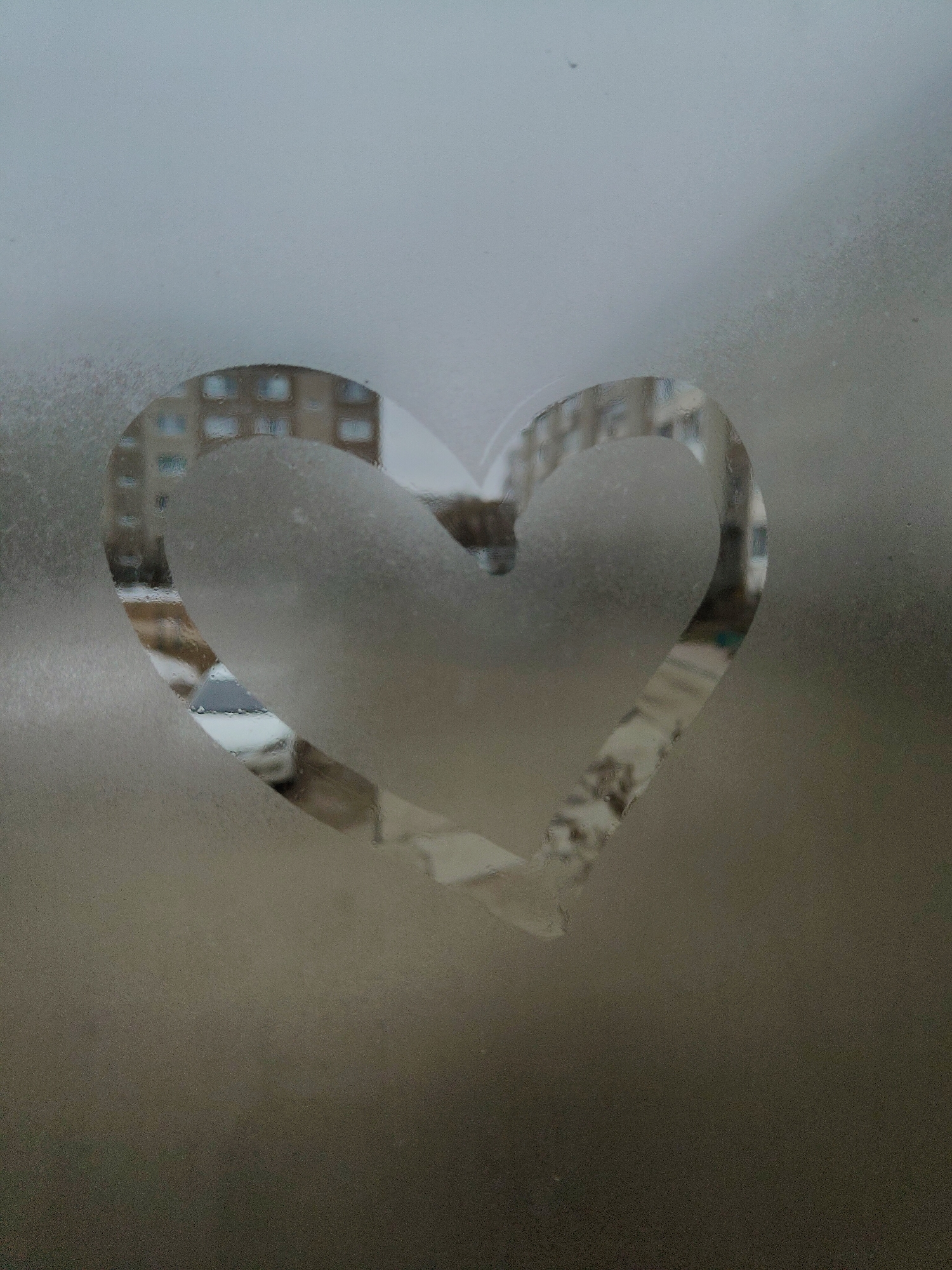 A heart shape is drawn into the condensation on a window. Outside the window, out of focus, is a cityscape. A building with multiple windows is visible. A car is also seen on the street. The rest of the window is covered in condensation.