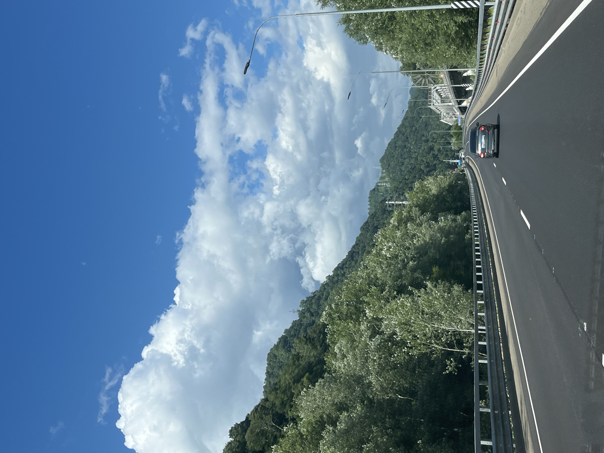 An eye-level, forward-facing shot captures a roadway in Kepsha, Russia, under a bright blue sky dotted with white clouds. The highway has two lanes in each direction, separated by a median, and a metal guardrail lines the outer edges. Green trees and densely wooded mountains flank the left side of the road, while a bridge and more trees are visible on the right. A silver car is in the right lane of the oncoming traffic, and other vehicles are further ahead on the road. Streetlights are positioned along the highway.