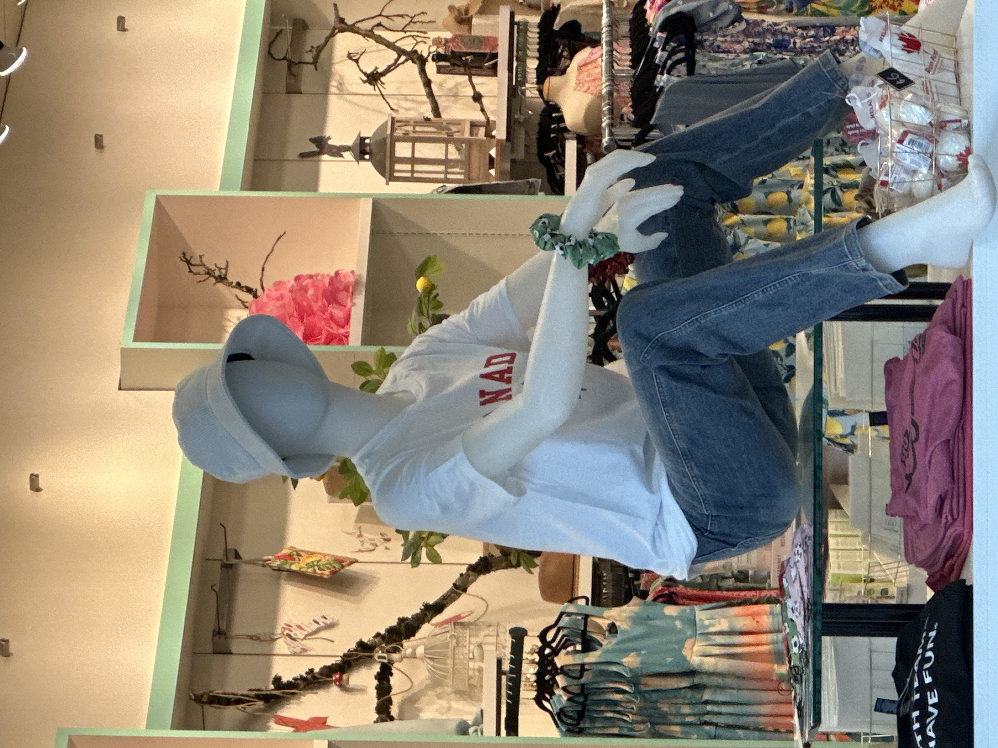 This image depicts a store display featuring a mannequin dressed in a white t-shirt with red lettering, blue jeans, a light blue bucket hat, and white socks. The mannequin is seated on a glass shelf in a retail environment. Behind the mannequin, the store displays clothing racks with various garments, decorative items such as a lantern and branches, and shelving units. On the shelf below the mannequin, several t-shirts are folded, and a wire basket contains small white objects with red maple leaf emblems and tags. The scene appears to be set in a retail store, likely a clothing boutique. The presence of maple leaf imagery and the lettering on the mannequin's shirt suggest a Canadian theme, potentially indicating the location is in Canada. Without further context or identifiable landmarks, a specific location within Burnaby, Canada cannot be determined from the image alone.