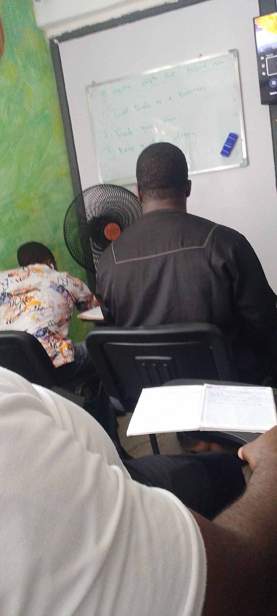 The image depicts a classroom setting in Lokoja, Nigeria. The view is from behind several students, most of whom are in focus.
In the background, a whiteboard displays the heading "#3 mental shifts that helped me" followed by a numbered list: "1) Treat trade as a Business," "2) Trade your plan," and "3) Keep a... diary." A blue eraser is positioned on the lower right corner of the whiteboard. A green wall with splotchy designs is visible to the left of the whiteboard. Adjacent to the right edge of the whiteboard, there is a dark-colored electronic display.
In the foreground, there is a desk fan with an orange center. A man in a black shirt with a cream-colored outline on the back sits directly in front of the fan. Another individual, wearing a patterned shirt with white, orange, and blue elements, sits to the left of the man in black.
In the very foreground, the shoulder and arm of another individual wearing a white shirt are visible. This person has a notebook placed on a desk attached to their chair.
The scene suggests a lecture or presentation taking place in a learning environment.