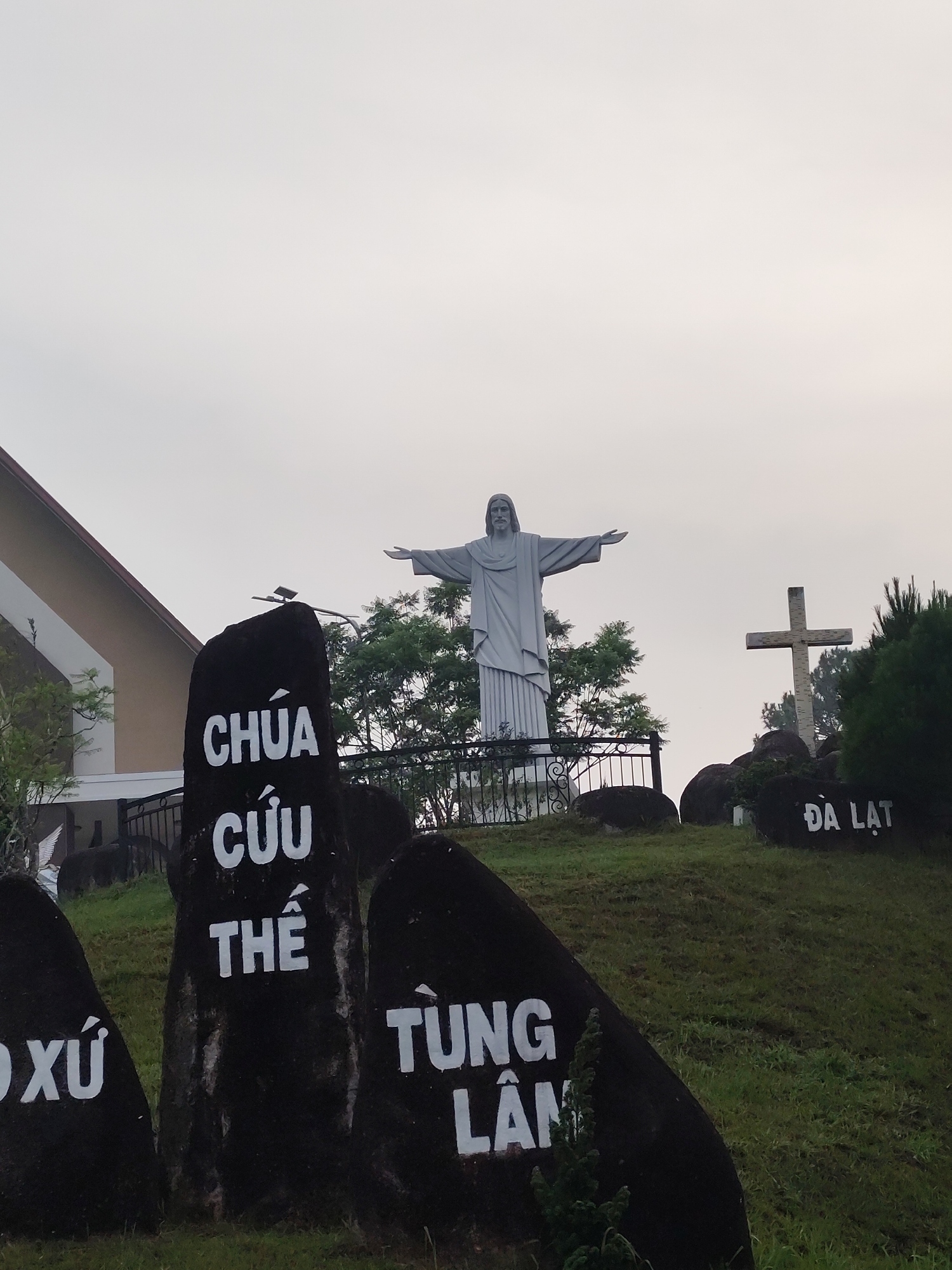 The image presents a serene religious landscape on an overcast day, likely in Da Lat, Vietnam, as indicated by the visible text. In the foreground, several large, dark, irregular-shaped rocks are scattered across a grassy slope. The central rock prominently displays the white Vietnamese text "CHÚA CỨU THẾ," which translates to "Savior Christ." To its right, another rock partially shows the text "TÙNG LÂM," and to the far left, a glimpse of "XỨ" is visible on another rock.
Dominating the midground atop the hill is a large, light grey statue of Jesus Christ, standing with his arms outstretched, reminiscent of the Christ the Redeemer statue. A black metal fence with decorative scrolls runs along the crest of the hill, just in front of the statue. Behind the Jesus statue, a cluster of green trees provides a natural backdrop. To the right of the statue, a tall, rugged wooden cross stands on a separate small mound. Below the cross, another dark rock clearly displays the white text "ĐÀ LẠT," confirming the city.
In the background to the left, part of a building with a light-colored wall and a reddish-brown roof is visible, suggesting a church or a related religious institution. The sky is uniformly light grey and overcast, contributing to a calm and contemplative atmosphere. The scene captures a prominent religious landmark, possibly a pilgrimage site, characterized by its statues, cross, and textual markers.