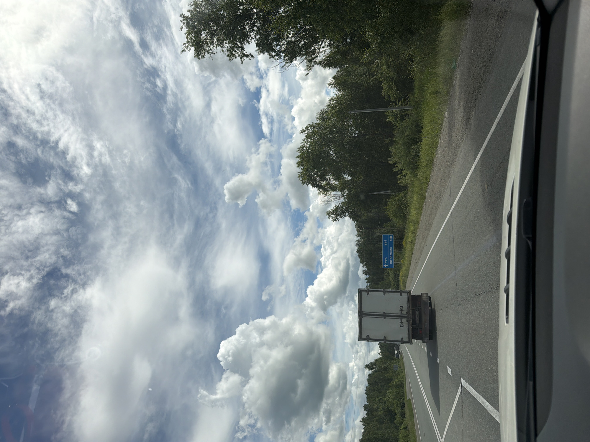 The image depicts a view from inside a vehicle, looking out at a two-lane asphalt road in a rural setting. A white truck with a closed trailer is driving in front of the vehicle. A blue road sign is visible on the right side of the road, indicating "УФА 446" and "ПИТОМНИК 1" with directional arrows. The road is flanked by dense green trees and vegetation on both sides, suggesting a natural landscape. The sky is partially cloudy with a mix of white cumulus clouds and thinner cirrus clouds against a blue sky. The overall scene suggests a journey on a highway in Russia, potentially near Poletaevo 1th based on the provided information, on a day with variable weather. There are no people visible in the image.