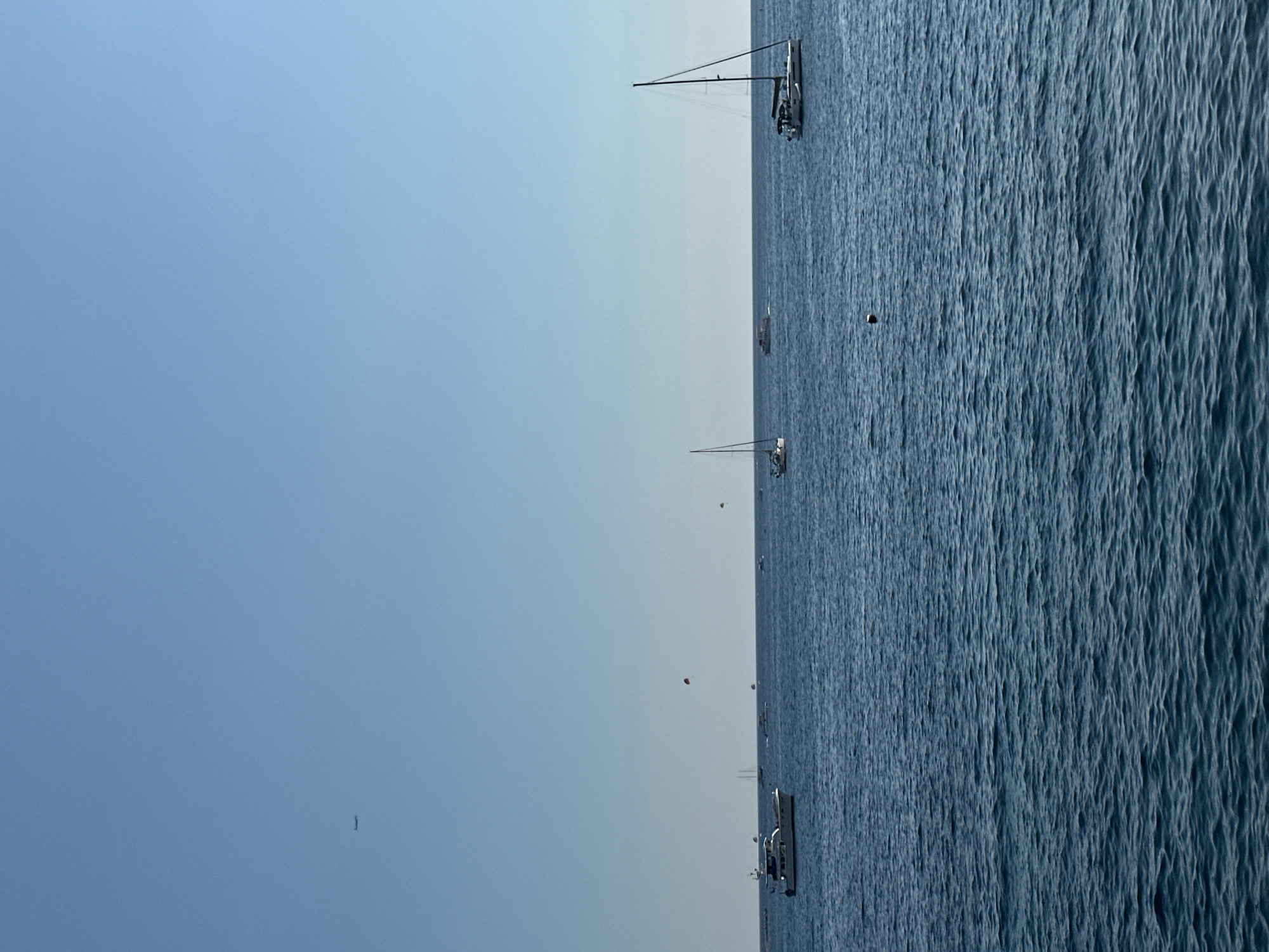 This image depicts a serene seascape with several boats scattered across the calm, blue water. The scene suggests a leisurely day on the sea, possibly in Budva, Montenegro, given the context. The sky is a clear, pale blue, indicating fair weather and likely daytime, possibly late morning or early afternoon given the gentle lighting.
The subjects in the image are primarily boats of various sizes and types. The most prominent is a sailboat with its mast and rigging clearly visible, carrying what appear to be a few people on board. Further out, other smaller boats and possibly a distant larger vessel can be seen. There are also a few small, spherical objects floating on the water, which could be buoys or markers. In the sky, a tiny dark speck could be a distant bird or an aircraft.
The overall activity is peaceful and leisurely, with the boats appearing to be either anchored or moving slowly. The gentle ripples on the water suggest minimal wind, contributing to the tranquil atmosphere. The location cues are limited to the maritime setting, but the clear skies and calm waters are conducive to outdoor recreational activities at sea. There are no discernible emotions conveyed, but the scene evokes a sense of calm and relaxation. No visible text is present in the image.