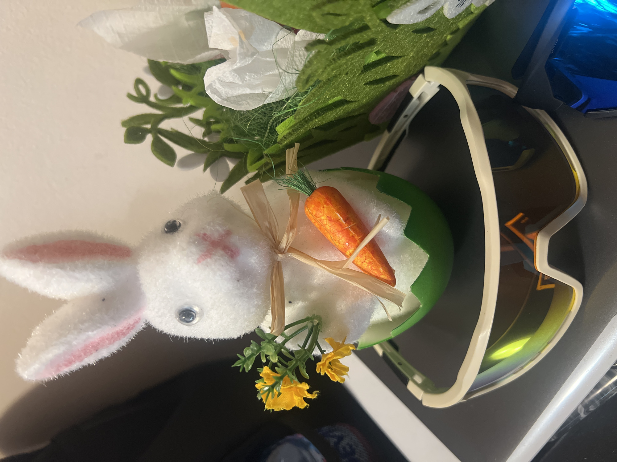 The image shows a close-up of a plush white bunny with pink-lined ears and a stitched pink "X" for a nose. The bunny is positioned next to a green egg-shaped container with a crack in the side, from which an orange carrot protrudes, held in place by a raffia ribbon. Yellow flowers are arranged near the base of the egg. Behind the bunny and egg is a mix of white paper flowers and green felt foliage. To the right of the egg and flowers, there's a pair of sunglasses with a cream-colored frame and dark lenses. The background is a dark surface with a hint of blue reflective material. There is no indication of location or city of Plovdiv, Bulgaria.