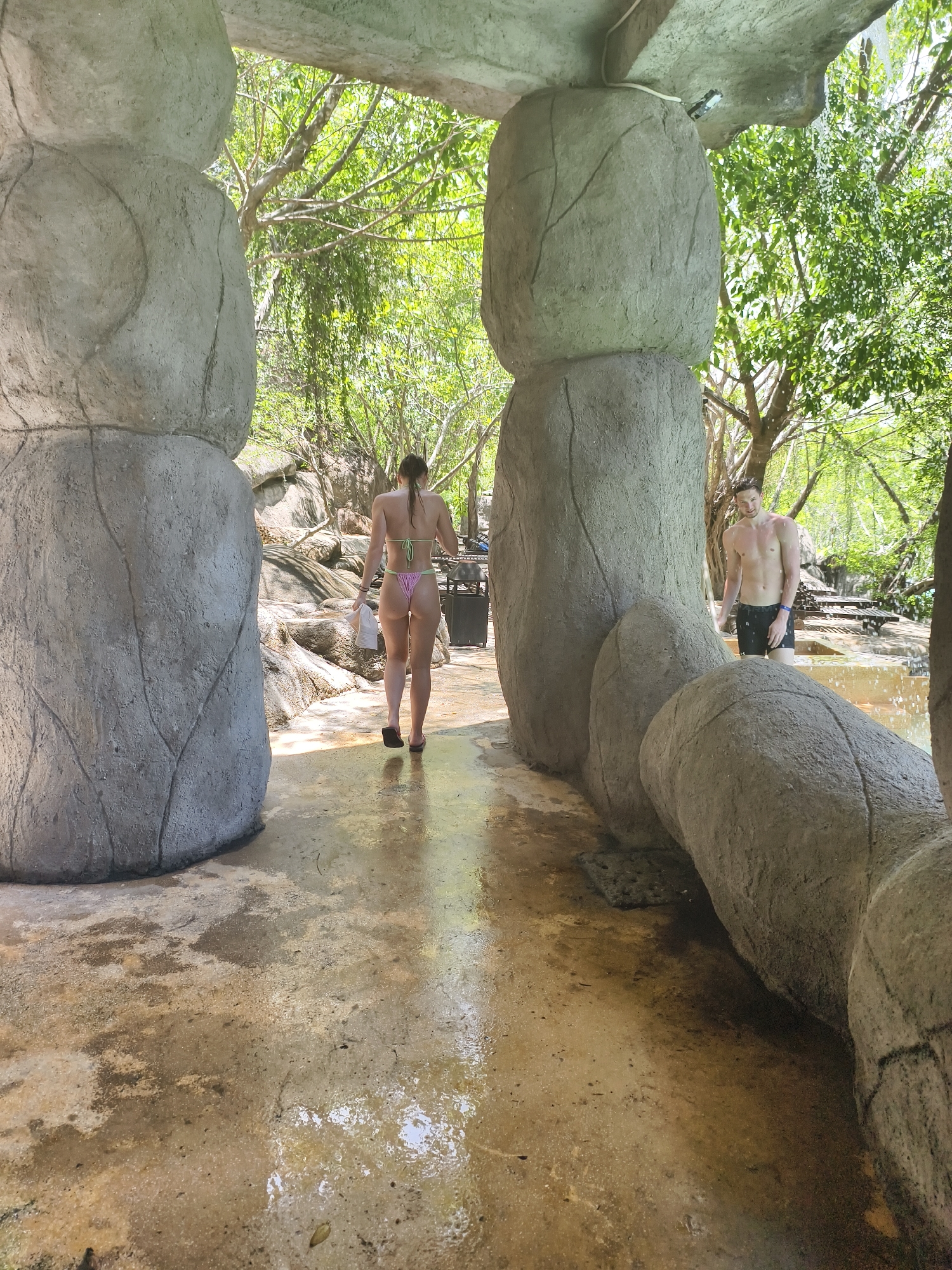 The image presents an outdoor scene in Tay Nha Trang Ward, Vietnam. Two individuals are visible: a woman with long brown hair wearing a string bikini walks away from the camera, carrying a white object in her left hand; a shirtless man in black swim trunks stands in a shallow pool of water, appearing to look towards the camera. The scene is framed by large, gray, artificially constructed rock formations that create an entranceway. Trees and greenery are visible in the background, adding to the natural setting. The ground is wet and appears to be made of concrete or stone.