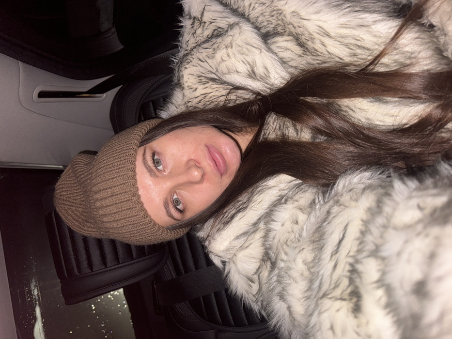 An image depicts a female individual seated within the dark interior of a vehicle, in what is contextually specified as the city of Высокиничи, Russia. The individual, positioned off-center, faces directly towards the camera, appearing to take a selfie. She wears a light brown ribbed knit beanie hat and a prominent light-colored, possibly faux fur, jacket with varying shades of white, grey, and dark brown tips. Her long, dark brown hair cascades over her shoulders and jacket. She has light-colored eyes, appearing green, and a neutral facial expression with full lips. Behind her, part of a black car seat headrest with horizontal stitching is visible to the left. The upper left portion of the image shows a dark window with indistinct reflections or precipitation, indicating an external low-light or nighttime environment. The overall lighting is dim, with the subject primarily illuminated from the front.