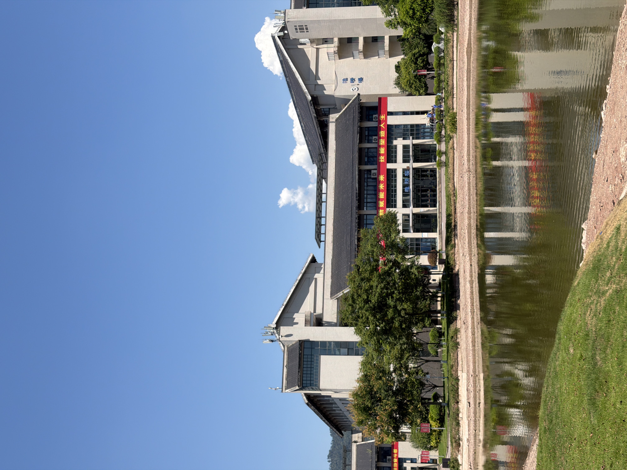 A wide-angle, eye-level shot captures a school building complex reflecting in a calm pond. The building is light-colored with dark tiled roofs and features multiple stories with many windows. Red banners with Chinese characters are visible on the building's facade, one of which reads "技能造人". Green trees are interspersed between the buildings and along the pond's edge. The pond's surface is rippled, creating a distorted reflection of the building and trees. The foreground shows a grassy bank sloping down to the pond, with some exposed soil and small rocks. The background consists of a clear, bright blue sky with a few white clouds, and a distant green hillside. The scene appears to be set in a park or campus environment within the city of Jiangjin, China.