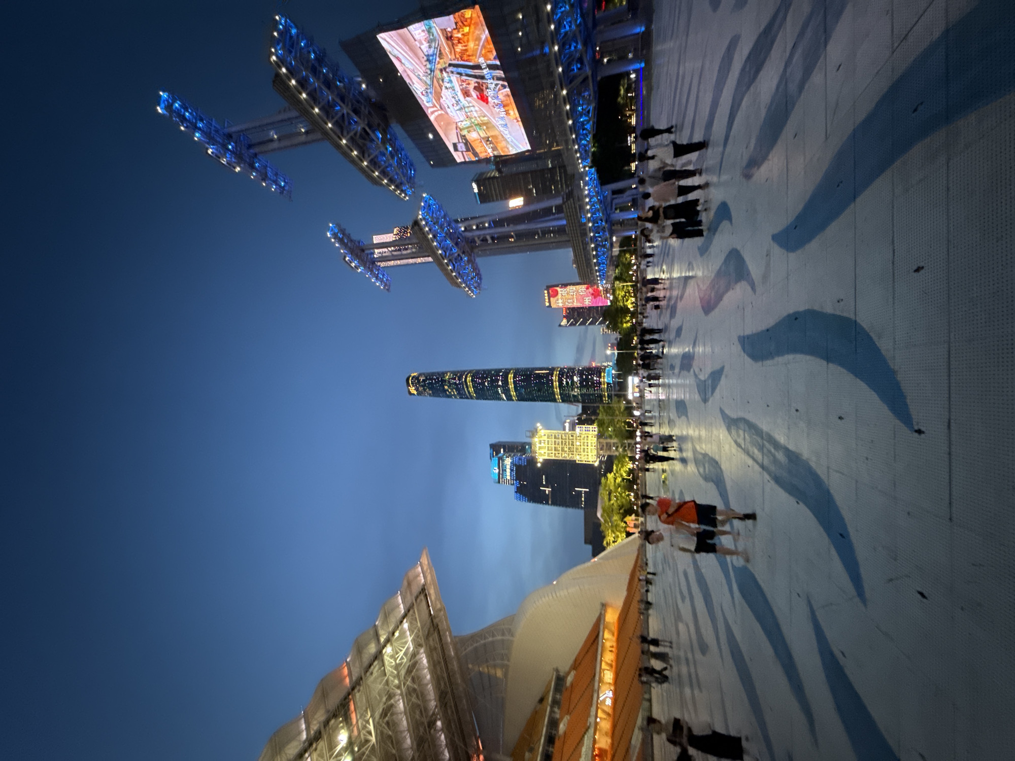 This image captures a wide-angle view of a plaza in Tianhe District, China, at dusk. The sky is a deep twilight blue, with a few wispy clouds. In the background, several modern skyscrapers are illuminated, including a prominent tall, slender building with lights tracing its outline. To the right, a large outdoor LED screen displays a colorful image, and above it, towering structures with blue and white lights resemble stadium lights.
In the foreground, a large, paved plaza features a repeating pattern of wide, curved blue shapes painted or embedded in the surface, creating a sense of movement and drawing the eye towards the city skyline. Several people are scattered across the plaza, walking and standing. Two men in casual attire are walking towards the viewer in the center of the frame, one wearing an orange t-shirt and shorts, the other a white t-shirt and shorts. Other individuals and small groups are visible in the mid-ground and background, contributing to the atmosphere of a public gathering space. To the left, a large, modern architectural structure with a sloped, illuminated roof and orange-colored seating or panels is partially visible. The overall scene depicts an urban environment at night, with a blend of modern architecture, public art on the ground, and people enjoying the open space.