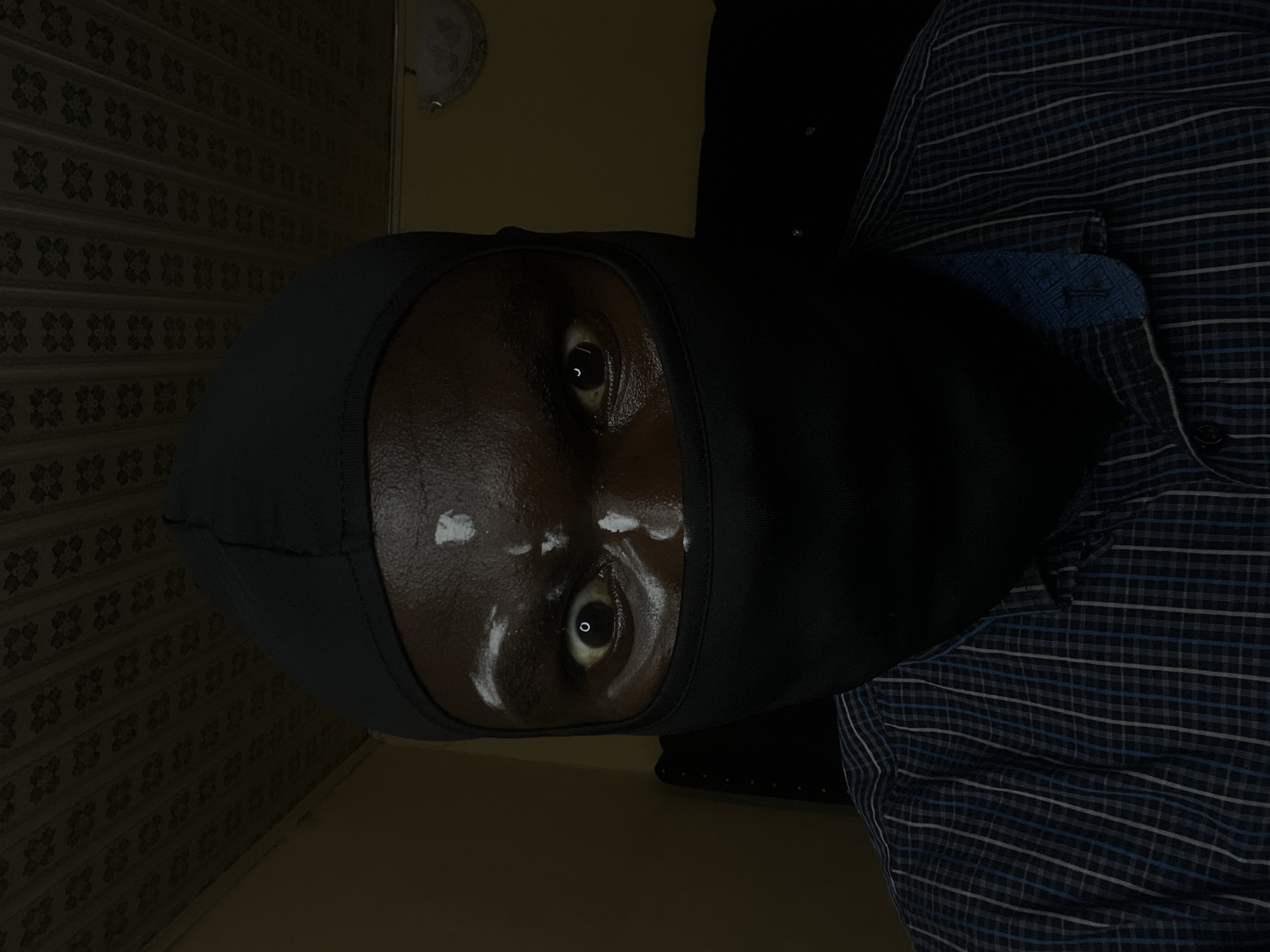 A person wearing a dark gray balaclava with streaks of white substance smeared on their forehead and cheeks is seen indoors. The individual is also wearing a blue plaid shirt with a buttoned cuff visible. The background includes a wall with a patterned design and a ceiling fixture. No specific location within Gura Top, Nigeria, can be determined from the image.