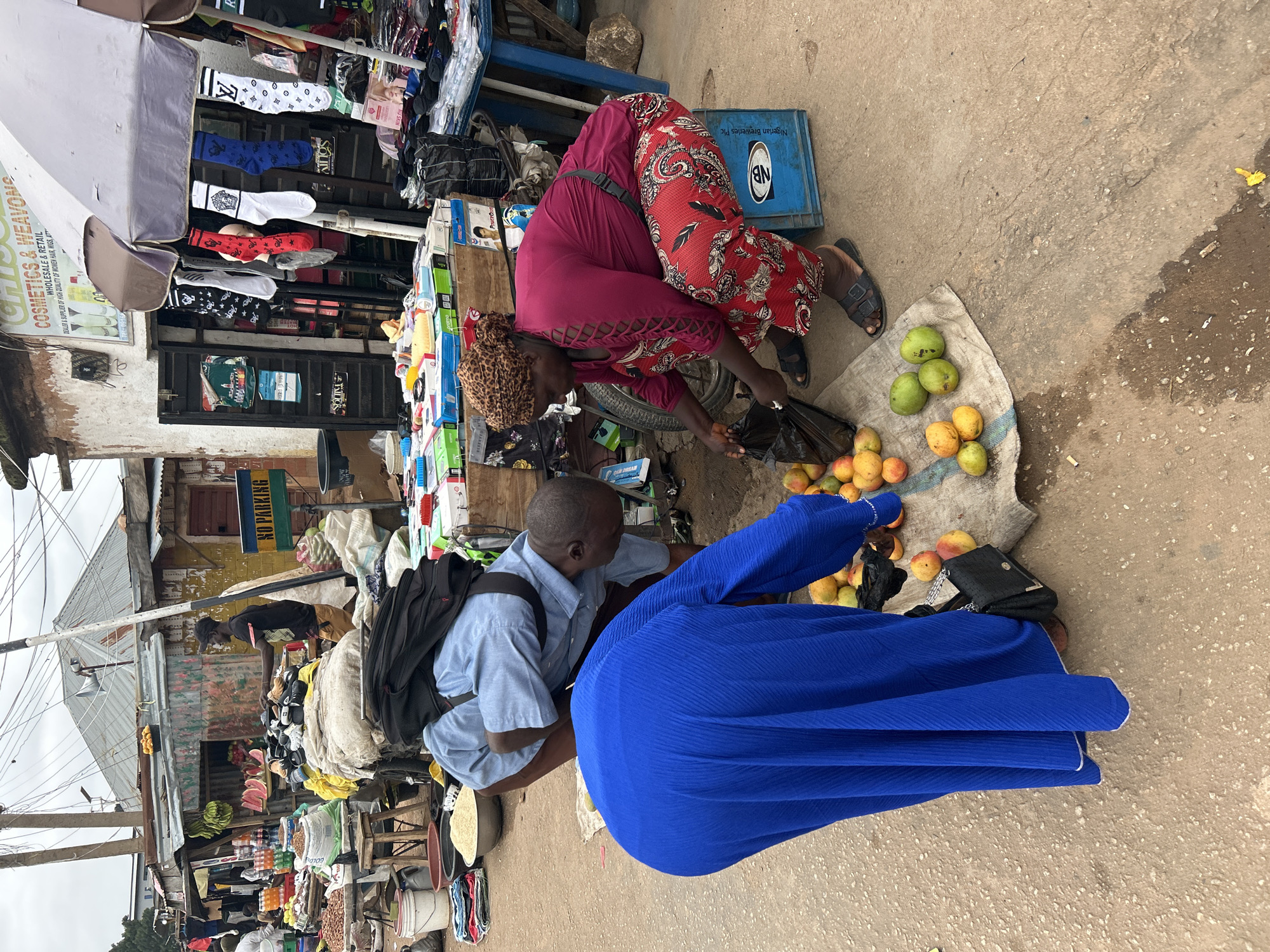 This image captures a vibrant street market scene in Jos, Nigeria, featuring two individuals engaged in commerce. In the foreground, a woman wearing a maroon top and red patterned trousers, with a leopard print head covering, is seen crouching. She is selecting items, likely mangoes, from a burlap sack on the ground and placing them into a black plastic bag. Nearby, a man in a light blue collared shirt and black backpack is also crouched, appearing to assist or interact with the woman. A collection of green, yellow, and reddish mangoes are spread on the sack.
The setting is an active market street lined with various stalls. Overhead, an umbrella canopy provides shade. A blue plastic crate bearing the text "Nigerian Breweries Plc" and the "NB" logo is visible near the woman. One stall displays an assortment of socks with different patterns, including what appears to be a Louis Vuitton pattern, and other items. Another stall advertises "COSMETICS & WEAVONS WHOLESALE & RETAIL DEALER & SUPPLIER OF HIGH QUALITY OF WOMEN HAIR, WIGS, AT" with a partial phone number "TEL:0916". Further down the bustling street, additional market stalls offer fresh produce like bananas and watermelon, and bags of what look like grains or seeds. A "NO PARKING" sign is also visible amidst the informal structures and overhead electrical wires. The ground is a mix of concrete and dirt, indicative of an outdoor market environment. The scene unfolds during daylight hours, under what appears to be an overcast sky or in shade, with no strong emotions discernible from the subjects.