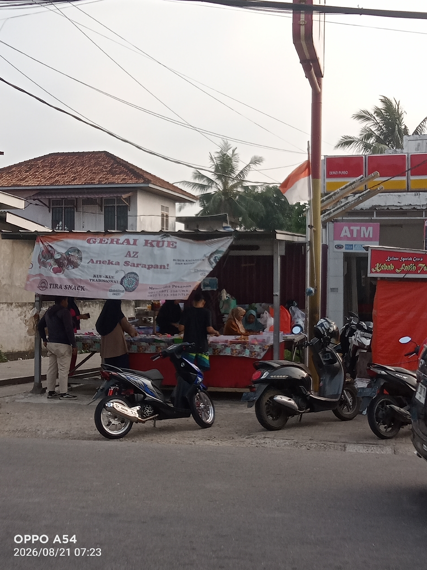 The image depicts a street scene in Jambi, Indonesia, likely in front of a small shop or food stall. In the foreground, two motorcycles are parked on the side of the road. One is a blue and white scooter with a prominent silver exhaust, and the other is a black scooter. The road surface appears to be asphalt.
Behind the motorcycles, a banner is displayed with the text "GERAI KUE AZ Aneka Sarapan!" which translates to "AZ Cake Stall Assorted Breakfast!". This suggests a food vendor selling cakes and breakfast items. Several people are visible behind the stall, some interacting with the items on display and others standing. The stall itself is a simple structure with a patterned tablecloth.
Further in the background, a building with a tiled roof is visible, along with palm trees and electrical wires overhead. A red and white Indonesian flag is flying from a pole. To the right, there is a sign with "ATM" in large letters, indicating the presence of an ATM machine, likely associated with a small convenience store or business. Another sign reads "Kuliner Syariah Grosir Kebah Avanti Juto". The overall impression is a bustling local street scene with a focus on food vending and everyday commerce.