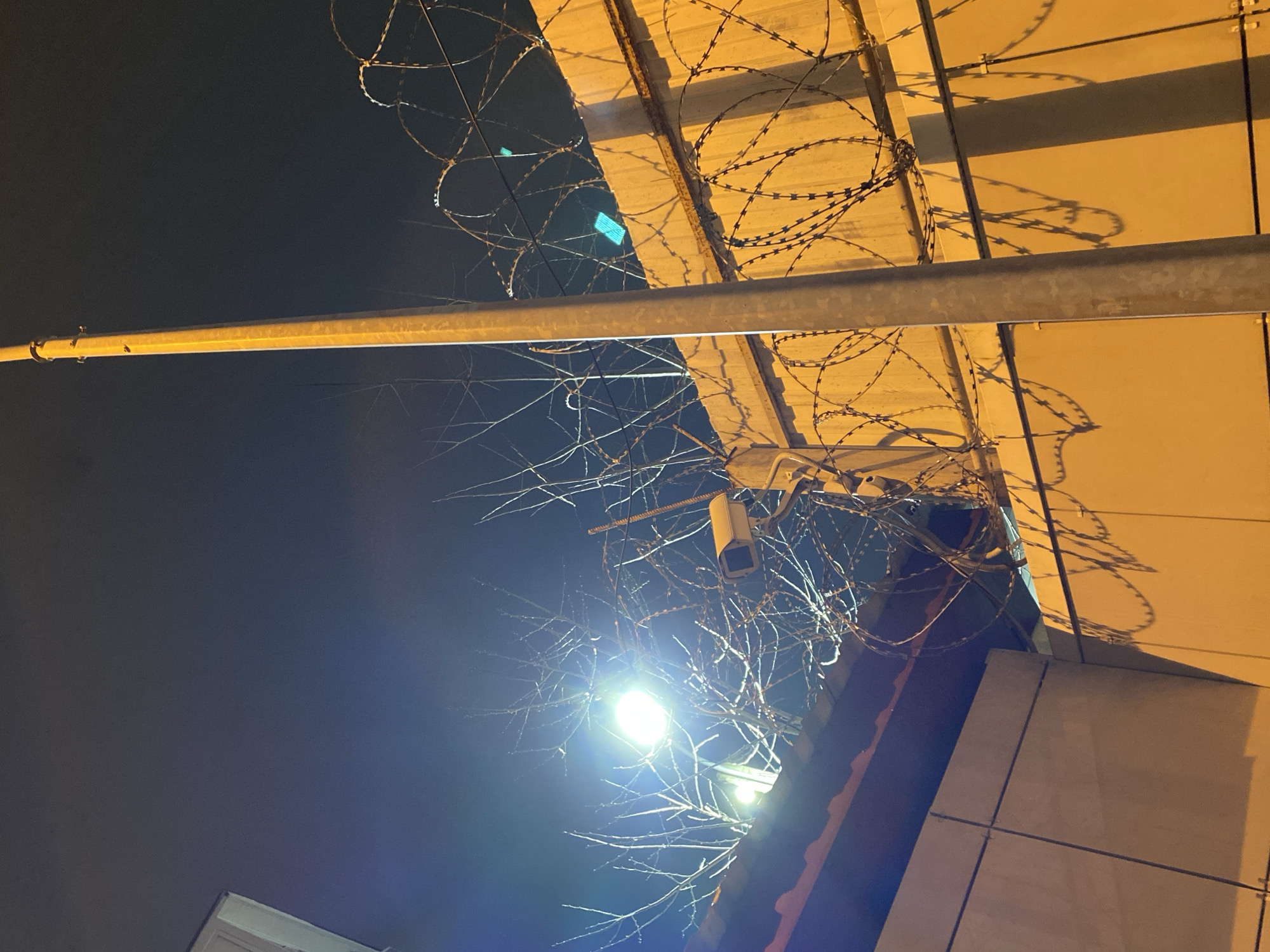 The image is an upward-looking shot taken at night. The sky is black and visible in the upper left and lower right corners. The primary subjects of the image include architectural elements such as the roof and walls of a building and barbed wire fencing. There are also leafless tree branches and a streetlight in the lower portion of the frame. A security camera is visible between the branches and below the barbed wire. The building is illuminated by artificial light, casting shadows from the barbed wire on the wall. The presence of barbed wire suggests the location may be a protected or restricted area. The location is Sochi, Russia.
