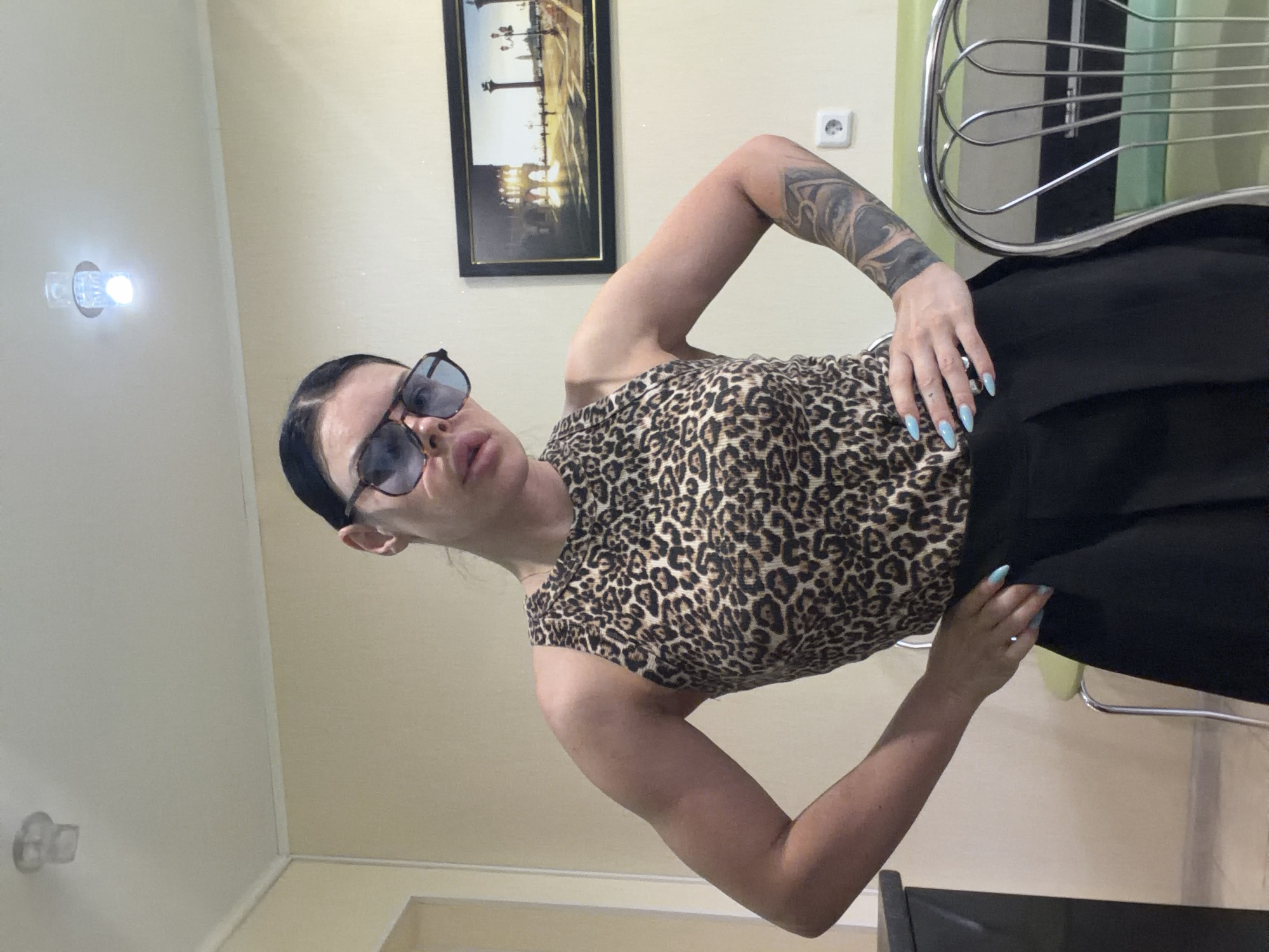 A woman stands in a room, posing with her hands on her hips. She is wearing a leopard print tank top, black pants, and sunglasses. She has dark hair pulled back and light blue nail polish. A framed picture hangs on the wall behind her, depicting a European cityscape. The room appears to be a residential interior, with a light-colored wall and ceiling. There is no visible indication of the specific location within Ufa, Russia.