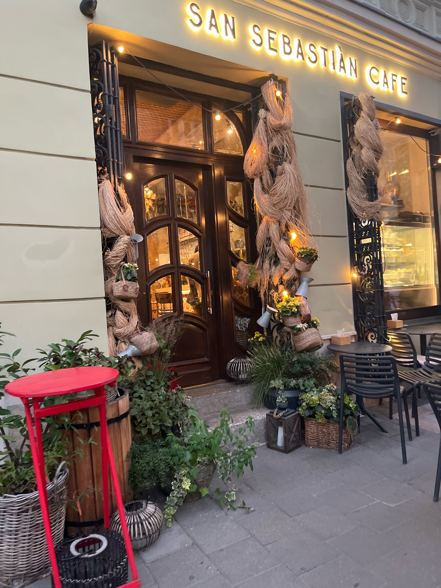 The image shows the facade of "SAN SEBASTIAN CAFE" in Lviv, Ukraine. The cafe features a dark wooden double door with arched glass panels, flanked by tall decorative arrangements of natural fibers. Small woven baskets with greenery and flowers are attached to these fiber decorations. The cafe's name is illuminated in bright yellow neon letters above the entrance. To the left of the door, a bright red stand holds a small round table, surrounded by potted plants in woven baskets and a wooden barrel. To the right of the entrance, an outdoor seating area is visible with dark metal tables and chairs. The pavement is made of gray stone tiles. The overall scene suggests a welcoming and artistically decorated entrance to a cafe.