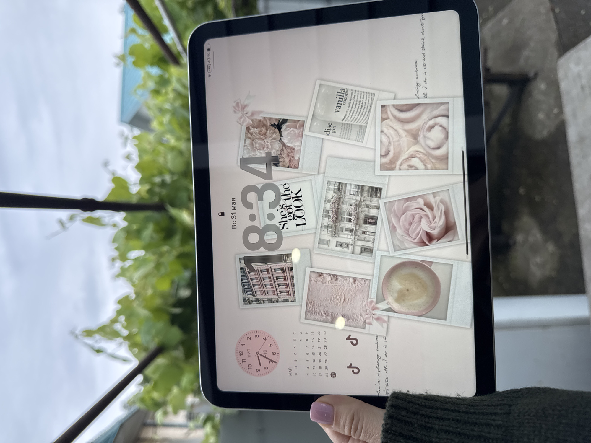 The image shows a hand holding a tablet displaying a customized lock screen with a light pink background. The lock screen features a collage of vintage-style images, including flowers, buildings, and a cup of coffee, arranged around the time "8:34" and the date "Bc 31 мая" (Wednesday, May 31st). A clock widget is visible near the bottom of the screen. The tablet's status bar indicates a VPN connection and a 43% battery level. Outside the tablet's frame, there's a blurred background showing foliage and what appears to be a building with a blue roof, possibly indicating an outdoor setting. The hand holding the tablet has a manicured nail with light purple polish. The edge of a green sweater is visible at the bottom corner. The scene appears to be in Sochi, Russia, as the date and time are displayed in Russian.