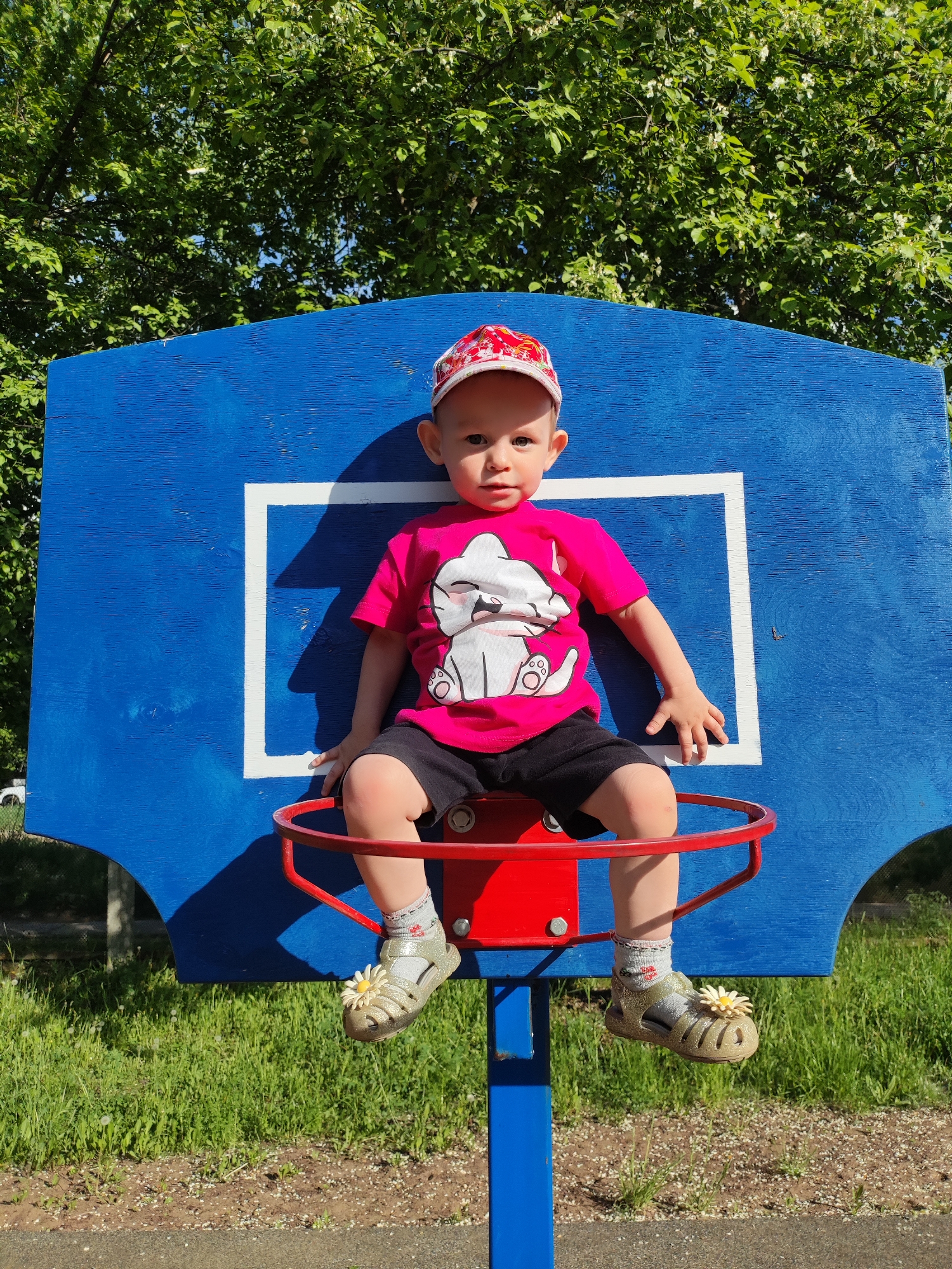 A toddler is sitting on a basketball hoop. He wears a red cap, a pink t-shirt with a cartoon cat, black shorts, and gold sandals adorned with flowers. He is sitting on the red basketball hoop that is connected to a blue backboard with a white square. The scene is set outdoors in a park in Chaikovsky, Russia with grass and trees in the background.