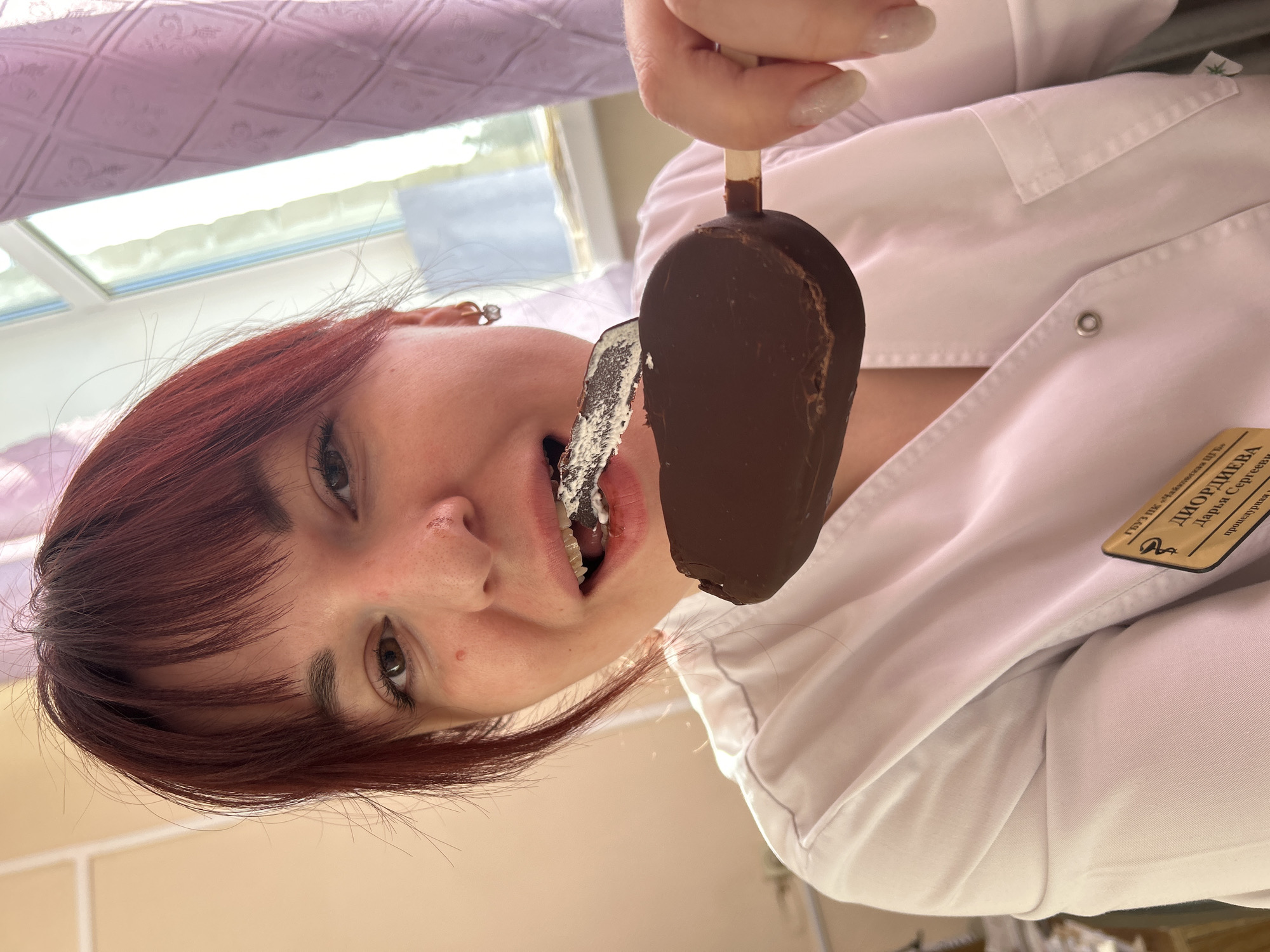 A woman with auburn hair is eating a chocolate-covered ice cream bar in Фоки, Russia. The woman is wearing a white lab coat with a name tag that reads "ДИОРДИЕВА Дарья Сергеевна процедурная м/с." The background is a window with a pink patterned curtain.