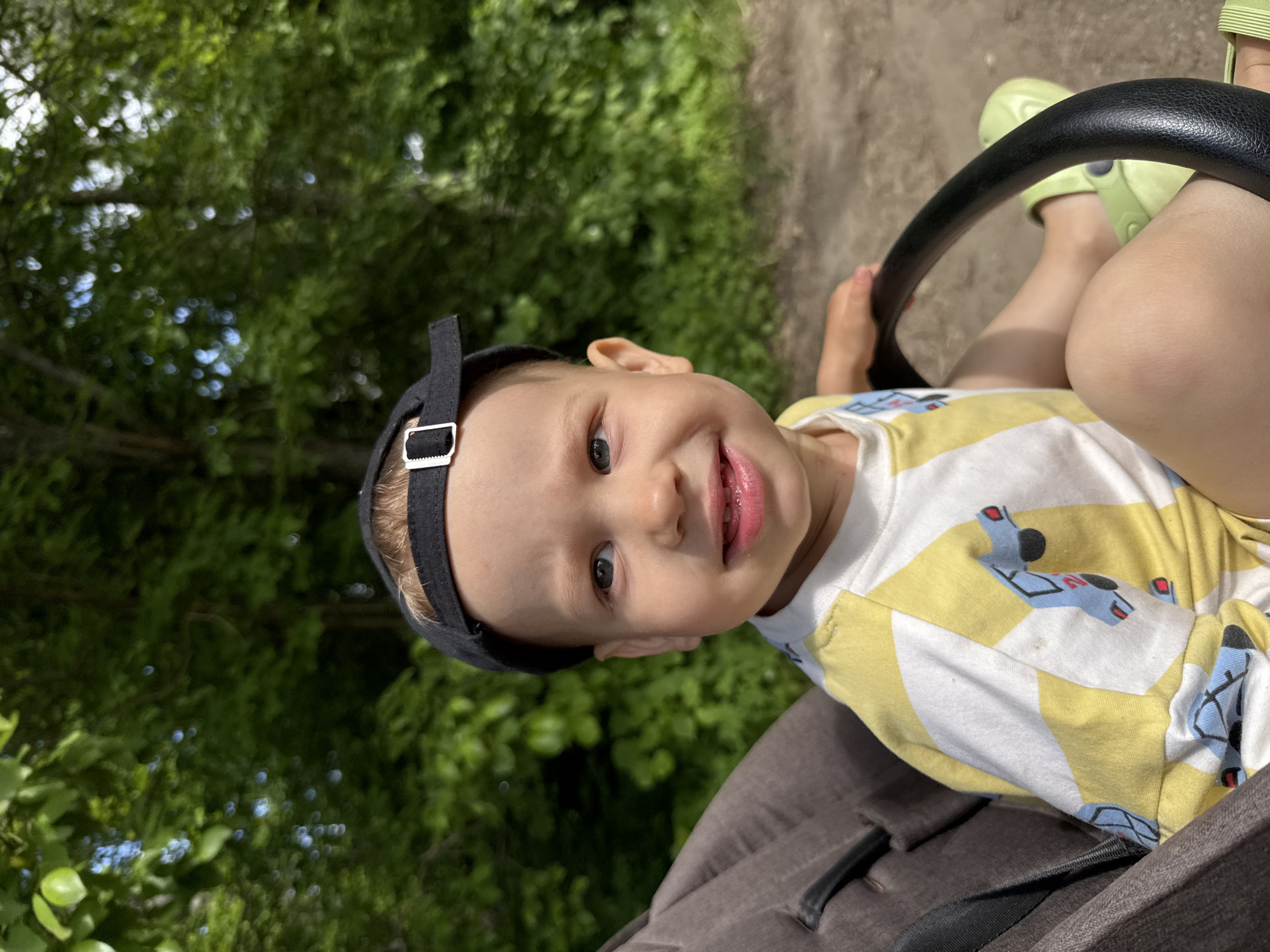The image captures a cheerful young child, likely a toddler, situated in what appears to be a stroller or carrier, outdoors in Rusanovo, Russia. The child is the primary subject, looking directly at the camera with a wide, open-mouthed smile that reveals a few teeth, conveying happiness and engagement. They are wearing a black baseball cap, reversed, and a yellow and white striped t-shirt adorned with blue and red patterns resembling stylized airplanes or vehicles. Light green clogs or sandals are visible on their feet. The child's right hand is gripping a black, textured bar, which is part of the stroller's frame.
The setting is a vibrant outdoor environment, dominated by lush green foliage from trees and bushes that create a natural backdrop. A light-colored dirt or gravel path is visible in the background, suggesting a walk or outing. The bright lighting indicates it is a sunny day. There is no visible text within the image.
