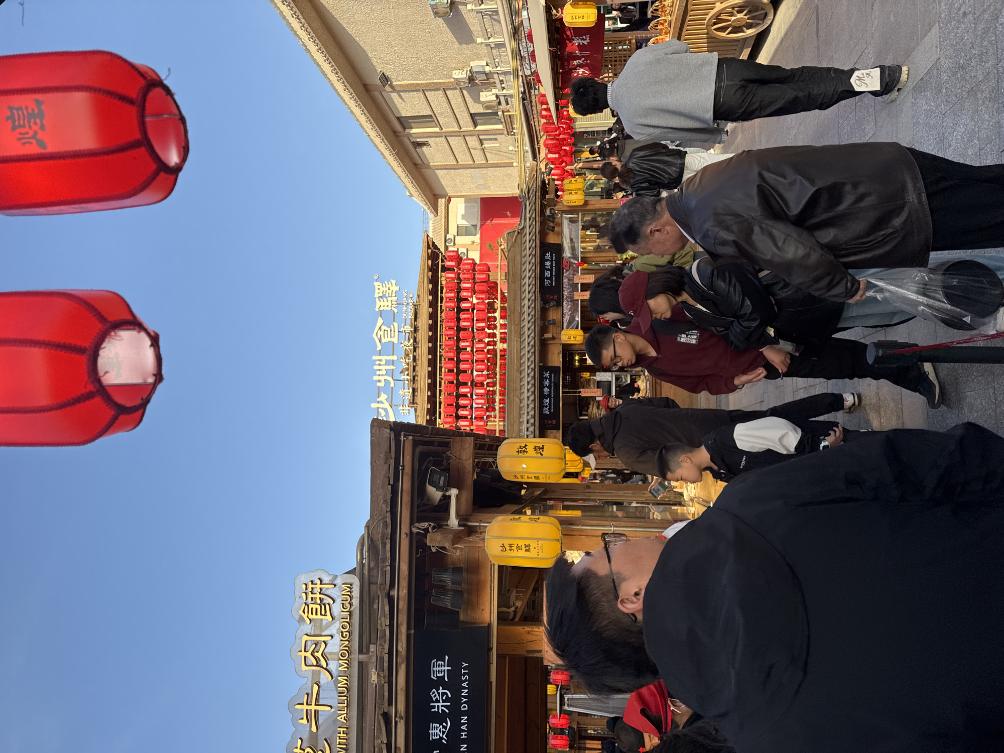 The image captures a bustling street scene in Dunhuang City, China. Numerous people of various ages stroll past shops and restaurants. Decorations include red and yellow lanterns. Signs are prominently displayed with Chinese characters alongside English translations. One restaurant's name is "Beef Pie With Allium Mongolicum." Other businesses reference the "Han Dynasty" and offer "Dunhuang Specialty Dishes."