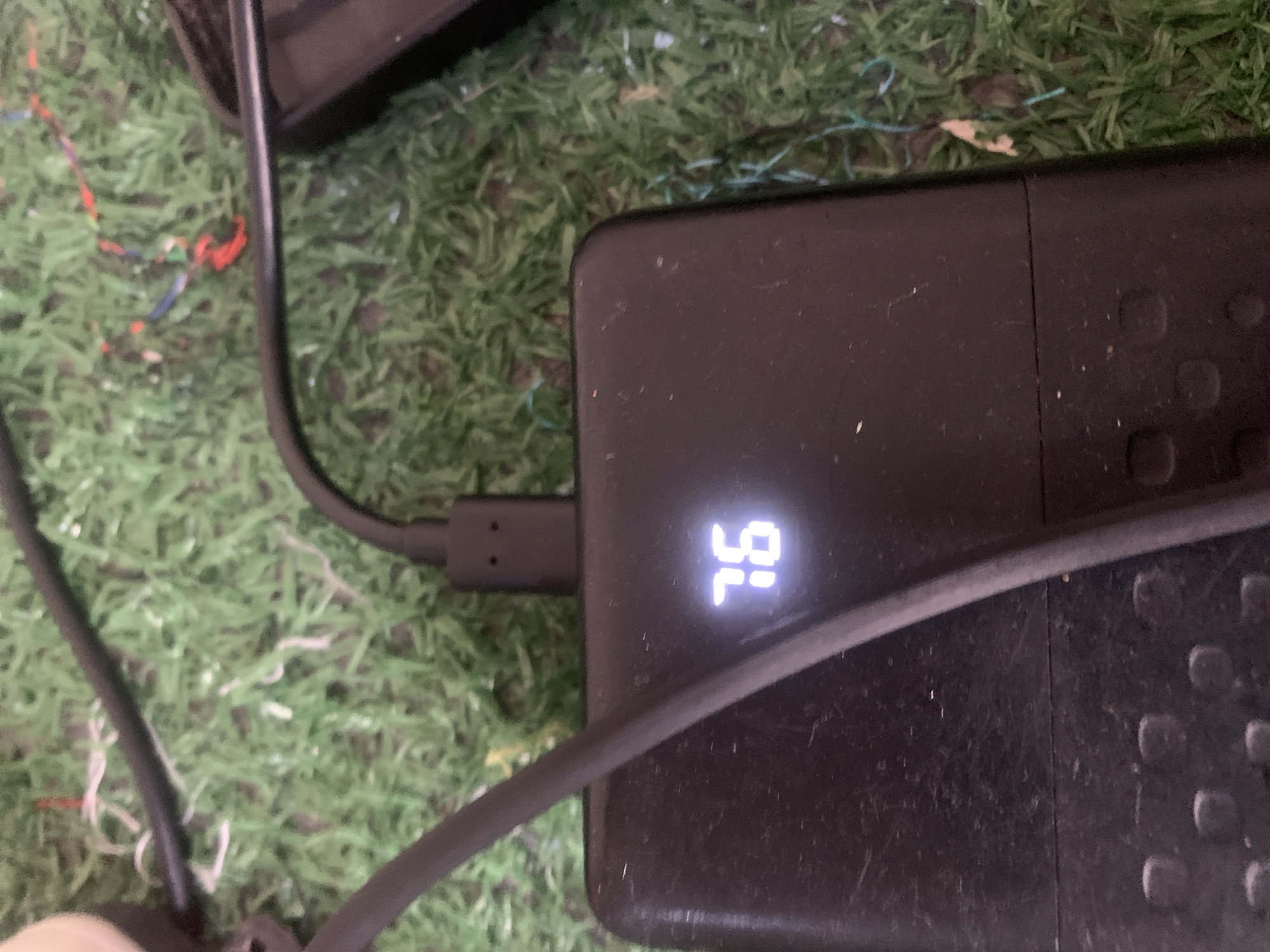 An outdoor, close-up, eye-level shot shows a black power bank on a green, artificial grass surface. The power bank has a digital display showing the number "76" in a bright, pixelated font. A dark grey USB cable is plugged into the top of the power bank and extends out of the frame to the left. A second, thicker black cable is also present, running diagonally from the top left to the middle right of the image. The background consists of the green artificial grass with some scattered, colorful wires, including red, blue, and white strands, partially visible. The overall scene is dusty and appears to be outdoors.