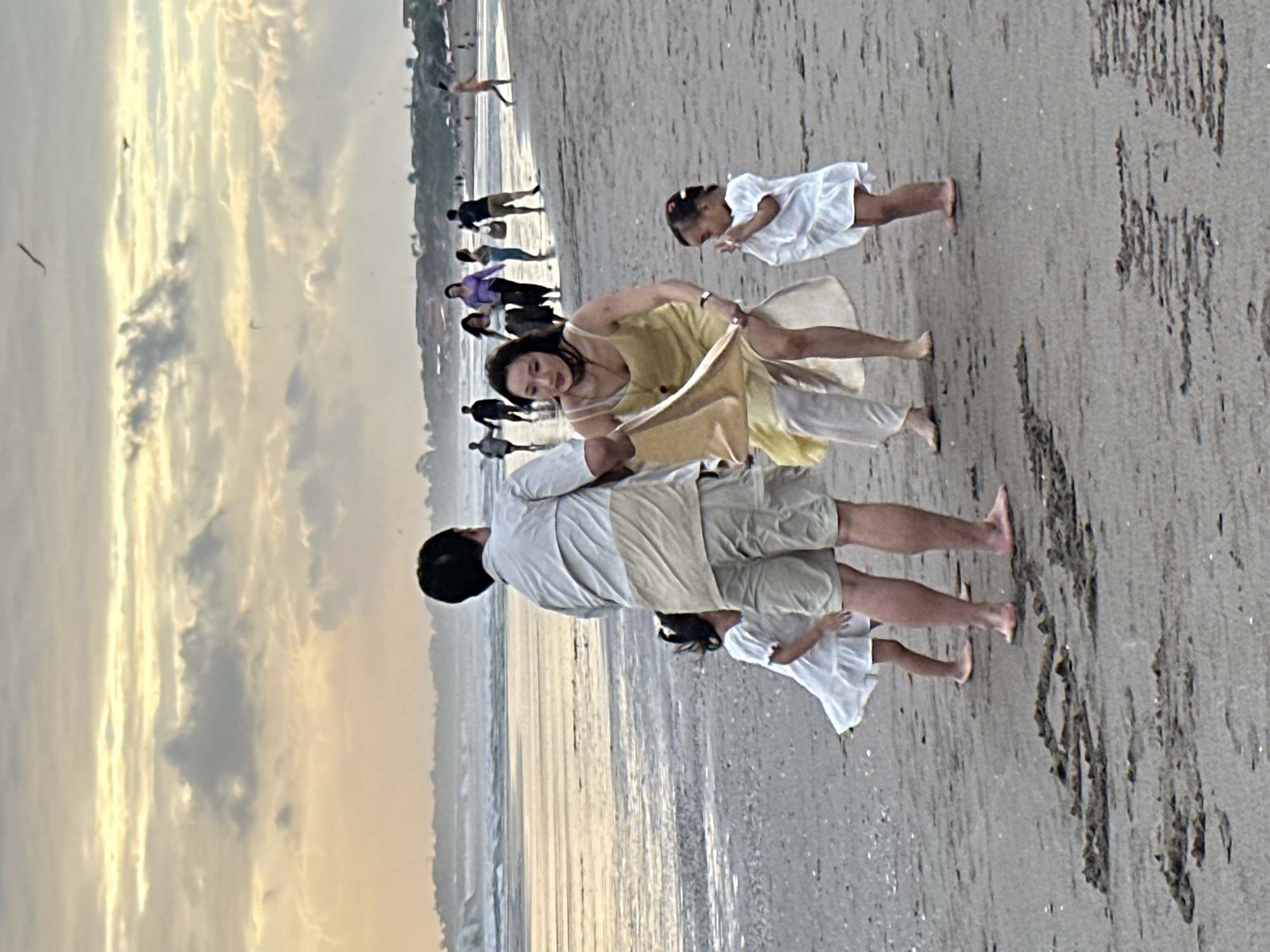 In North Kuta, Indonesia, a family is gathered on a beach with a body of water in the background. The sky above is filled with clouds, with a soft, yellowish light indicating it may be sunset or sunrise. In the foreground, a man wearing a light-colored short-sleeved shirt and beige shorts stands with his back to the camera. To his left, a young girl in a white dress is partially visible. In front of the man, a woman in a yellow dress and light-colored pants is holding a light-colored fabric. To her right, another young girl in a white dress stands. In the middle ground, several other people are walking along the beach, and in the background, there are more figures and structures, suggesting a populated area. The sand on the beach has some textures and markings.