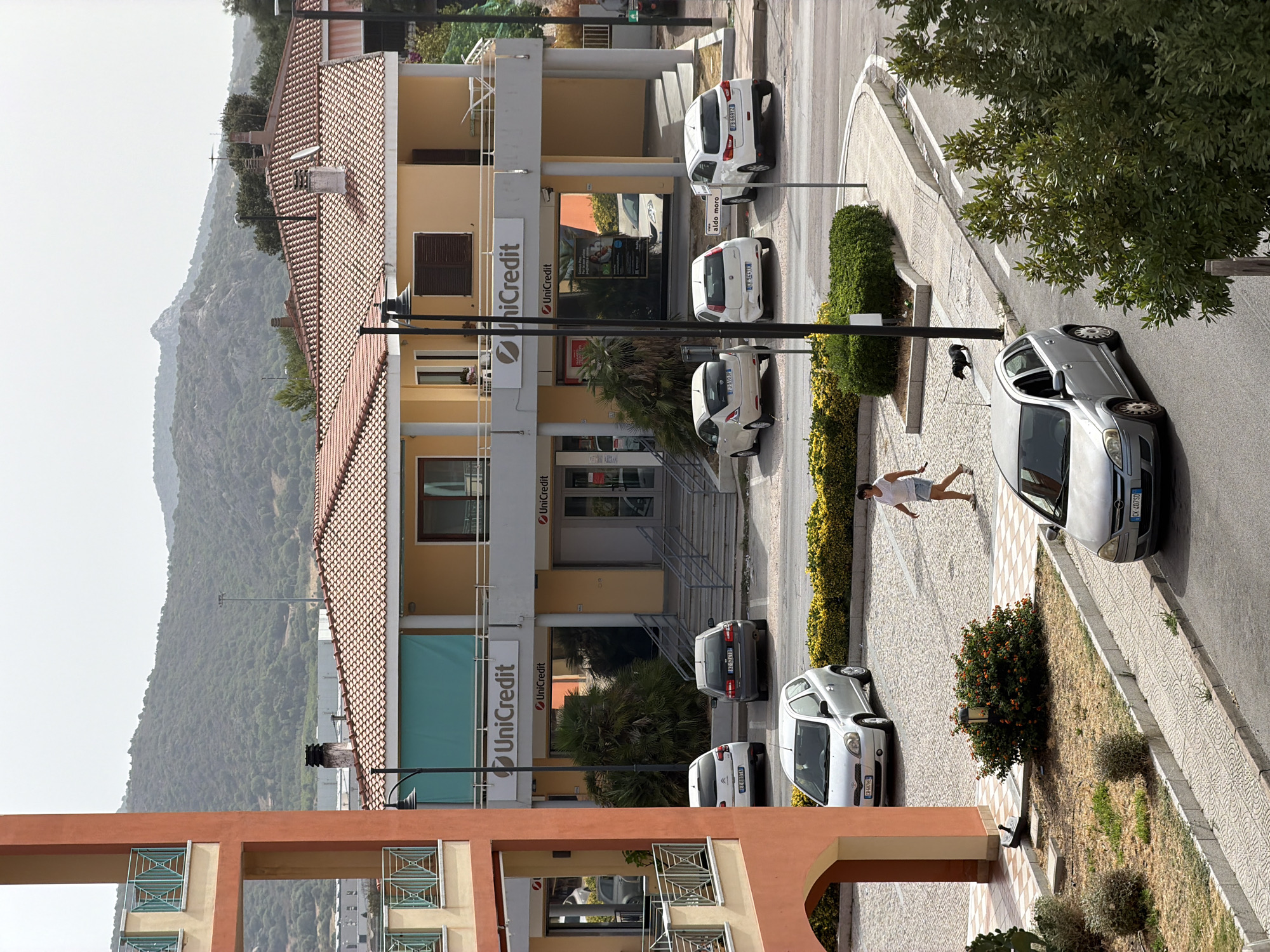 In Olbia, Italy, a woman in a white t-shirt and denim shorts walks down a cobblestone sidewalk. She is walking away from the camera, and a small black dog is trotting beside her. In the background, a row of cars is parked in front of a yellow building with a red-tiled roof. The building has several windows and signs for "UniCredit." A mountainous landscape is visible in the distance, shrouded in a light haze. The scene appears to be a quiet, daytime street in a Mediterranean town.