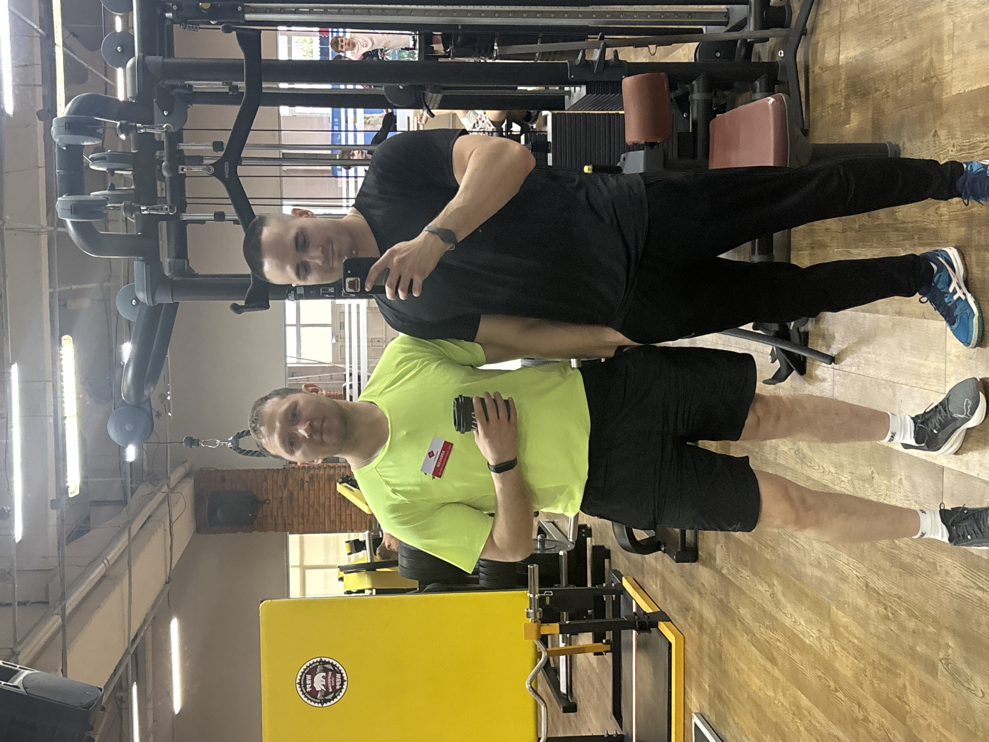 The image shows two men posing for a selfie in what appears to be a gym. One man, wearing a black t-shirt and black pants, is holding a phone and taking the picture. The other man, wearing a lime green t-shirt and black shorts, stands beside him. Gym equipment is visible in the background. A sticker with the inscription "Мебель Велесница Чайковский" (Furniture Velesnitsa Chaikovsky) is visible on a yellow panel, indicating the location is Chaikovsky, Russia.