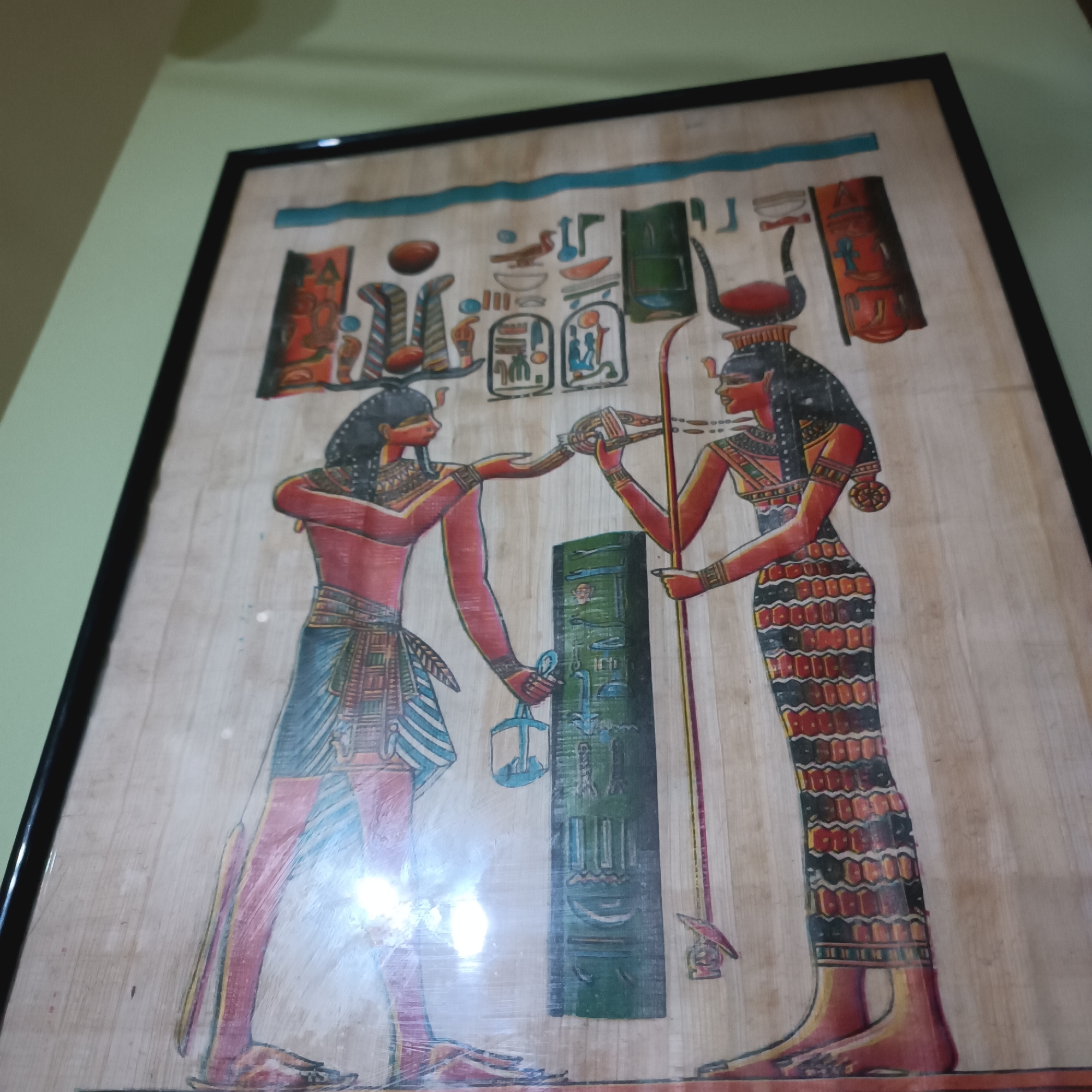 The media file displays a framed artwork, likely an ancient Egyptian-style painting on papyrus, featuring two prominent figures. The background material of the artwork is light-colored and fibrous, characteristic of papyrus, and it is enclosed within a simple black frame. Reflections are visible on the surface, suggesting it is under glass or a protective layer.
The central subjects are two figures depicted in the distinctive ancient Egyptian artistic convention, with profile heads and legs, and frontal torsos. The figure on the left appears male, with dark skin, a red upper body, and a blue and white striped kilt. He holds a blue, lantern-like object containing an ankh symbol in his left hand, while extending his right hand towards the second figure.
The figure on the right is female, also with dark skin, adorned with a long, intricately patterned dress in shades of red, black, and yellow. She wears a prominent headdress consisting of a sun disk cradled between cow horns, typically associated with deities such as Hathor or Isis. She holds a staff in her left hand and extends her right hand towards the male figure, seemingly engaged in an exchange or offering a serpentine object.
Between the two figures stands a tall, green, rectangular object, possibly a stele or column, covered with visible hieroglyphic markings. Additional Egyptian hieroglyphs and cartouches are depicted in the upper part of the artwork. The overall scene suggests a ritual, a divine interaction, or a symbolic presentation. The artwork utilizes a vibrant palette of red, blue, green, yellow, and black colors.