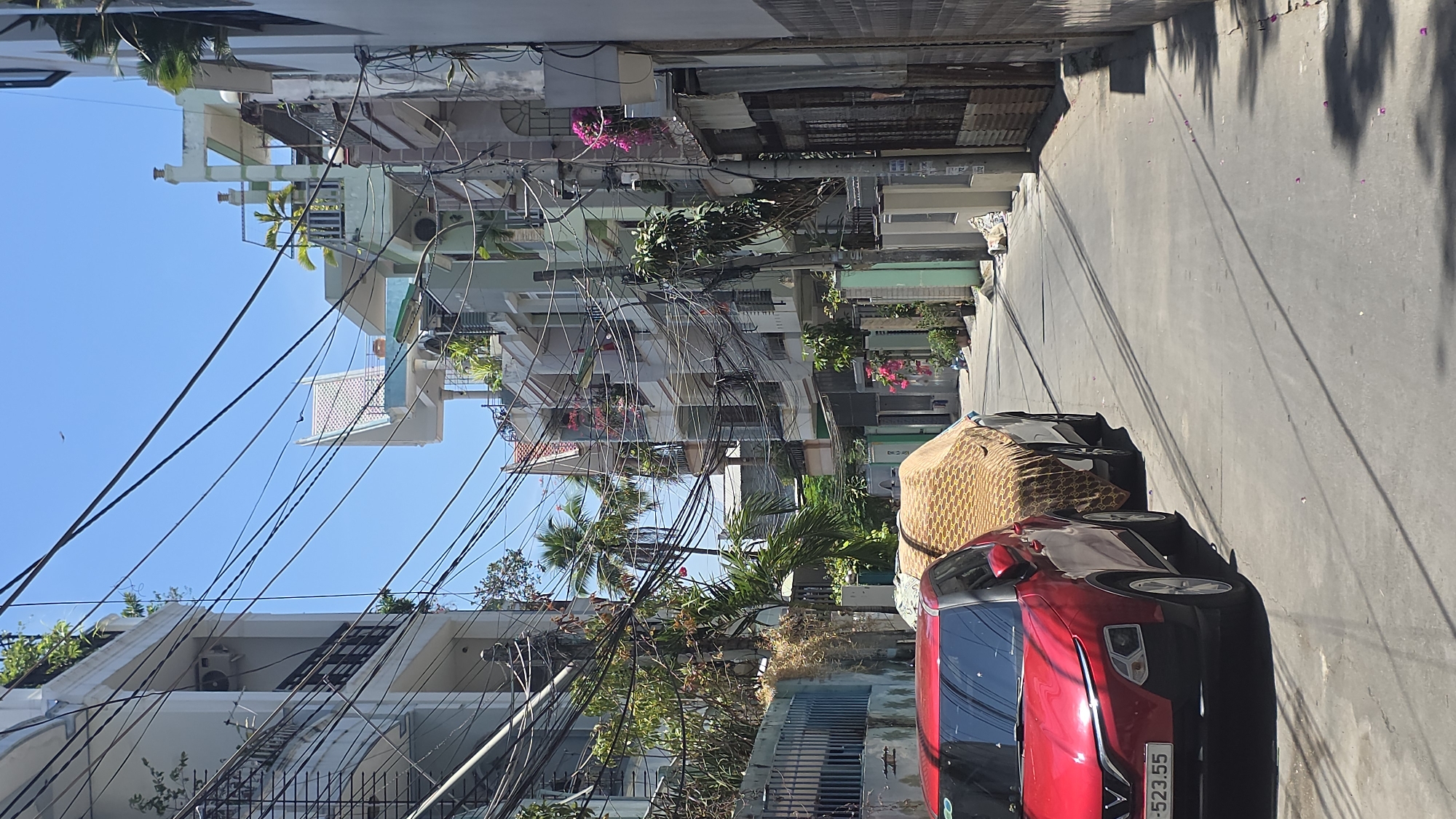 The image depicts a street scene in Nha Trang Ward, Vietnam, characterized by a narrow street lined with buildings. Overhead, a dense network of electrical wires crisscrosses the sky, adding to the visual clutter. A red car and another vehicle covered with a patterned cloth are parked on the side of the street. The buildings vary in height and architectural style, some adorned with plants and flowers. The street surface is gray, and shadows are cast by the buildings and trees. The overall impression is of a densely populated, urban environment.