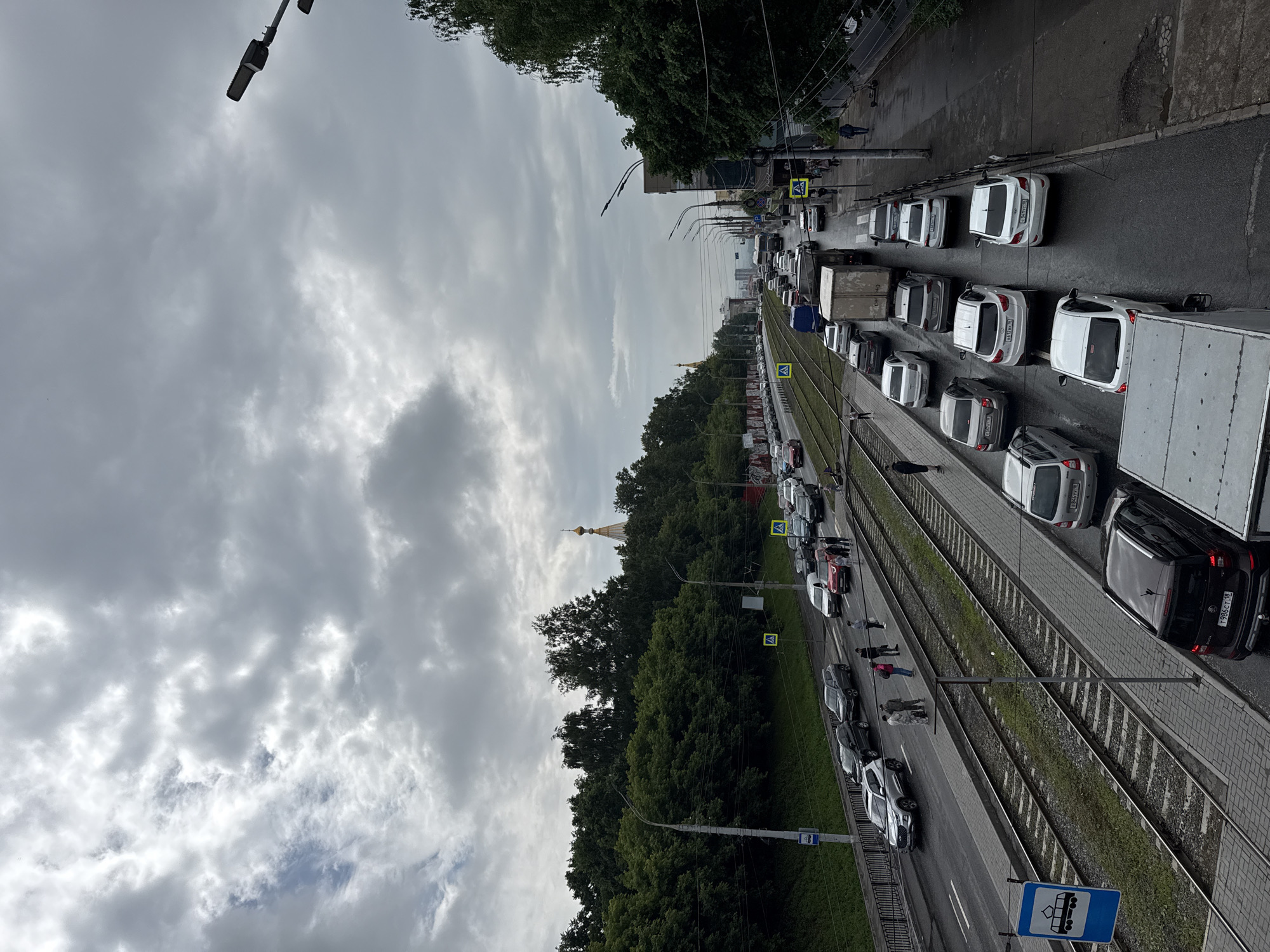 The image depicts a traffic jam on a cloudy day in Izhevsk, Russia. A wide street runs diagonally from the lower left to the upper right of the frame, with tram tracks running parallel to it on the left side. Cars are backed up in multiple lanes. On the left side of the street, there is a pedestrian walkway and a green embankment lined with trees, with a golden-domed church visible in the distance. On the right side, there is a sidewalk with more trees and buildings. Pedestrians are visible on the sidewalks and waiting at a crosswalk. One individual is operating a scooter.