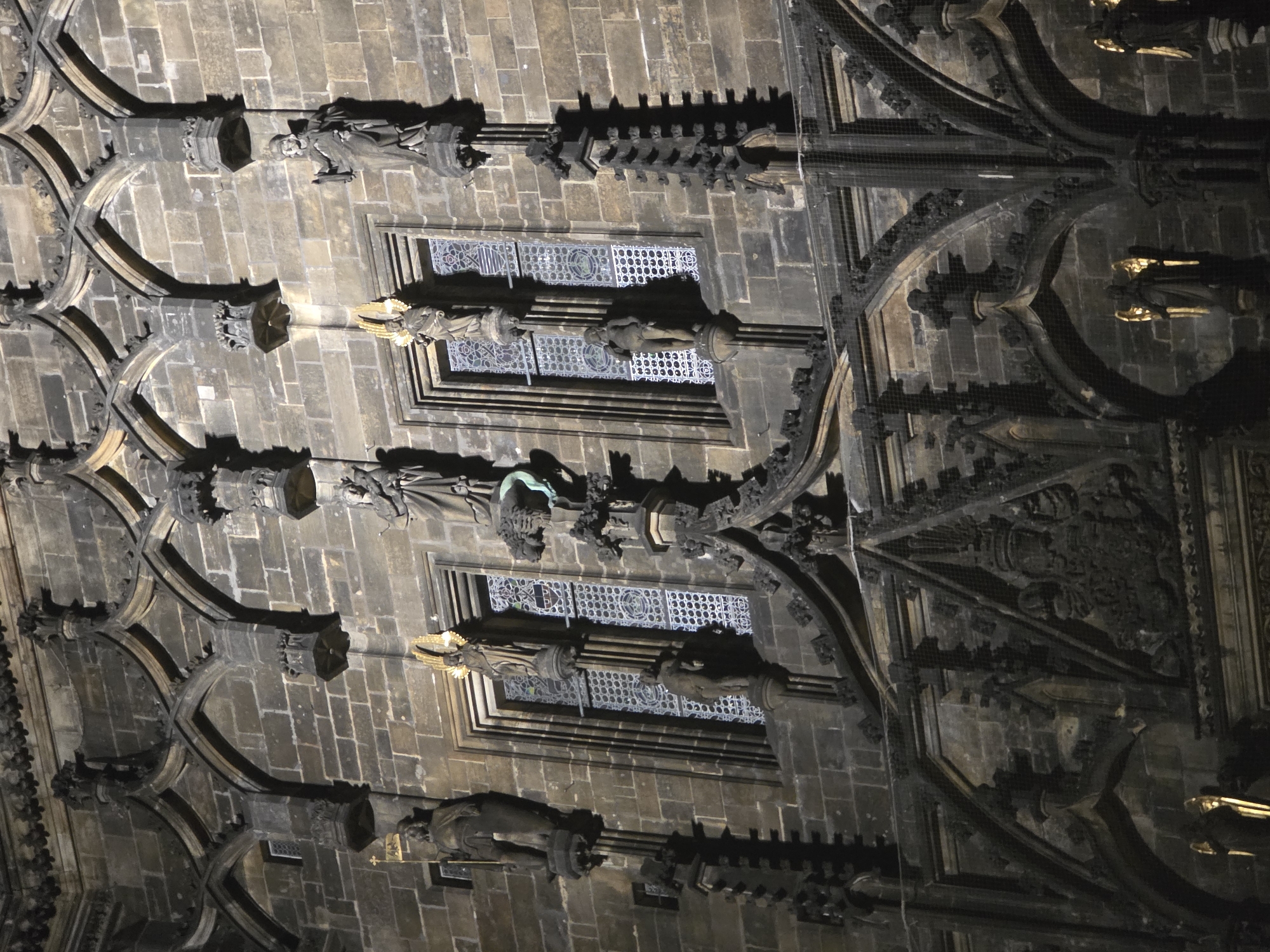 This image presents a dramatic, upward-looking view of an ornate architectural structure, likely a Gothic or Neo-Gothic ceiling or high wall within a grand building. The scene is dimly lit, suggesting nighttime or an interior setting, with artificial light dramatically illuminating specific features and casting deep shadows.
The primary subjects are intricate stone architecture and numerous statues. The architecture features complex arched structures, possibly rib vaults or detailed tracery, supported by decorated columns or pilasters. Windows with elaborate grilles or patterned glass are visible between these architectural elements, adding to the decorative complexity. The stone is predominantly dark gray and brown, indicating age and texture.
Throughout the structure, multiple carved human figures are positioned on pedestals or integrated into the architectural design. Many of these statues appear to be religious figures; one figure prominently displays bright golden wings and a halo, strongly suggesting an angel or saint. Another figure features a distinct green element, possibly part of its attire or an object it holds. The figures are intricately carved, showing detailed drapery and specific postures.
The artificial lighting enhances the texture and three-dimensionality of the carvings and statuary, creating a play of light and shadow that emphasizes the grandeur and historical depth of the structure. The overall impression is one of majestic scale, meticulous craftsmanship, and historical significance, typical of a cathedral, a large church, or a monumental public building. No visible text is present in the image.