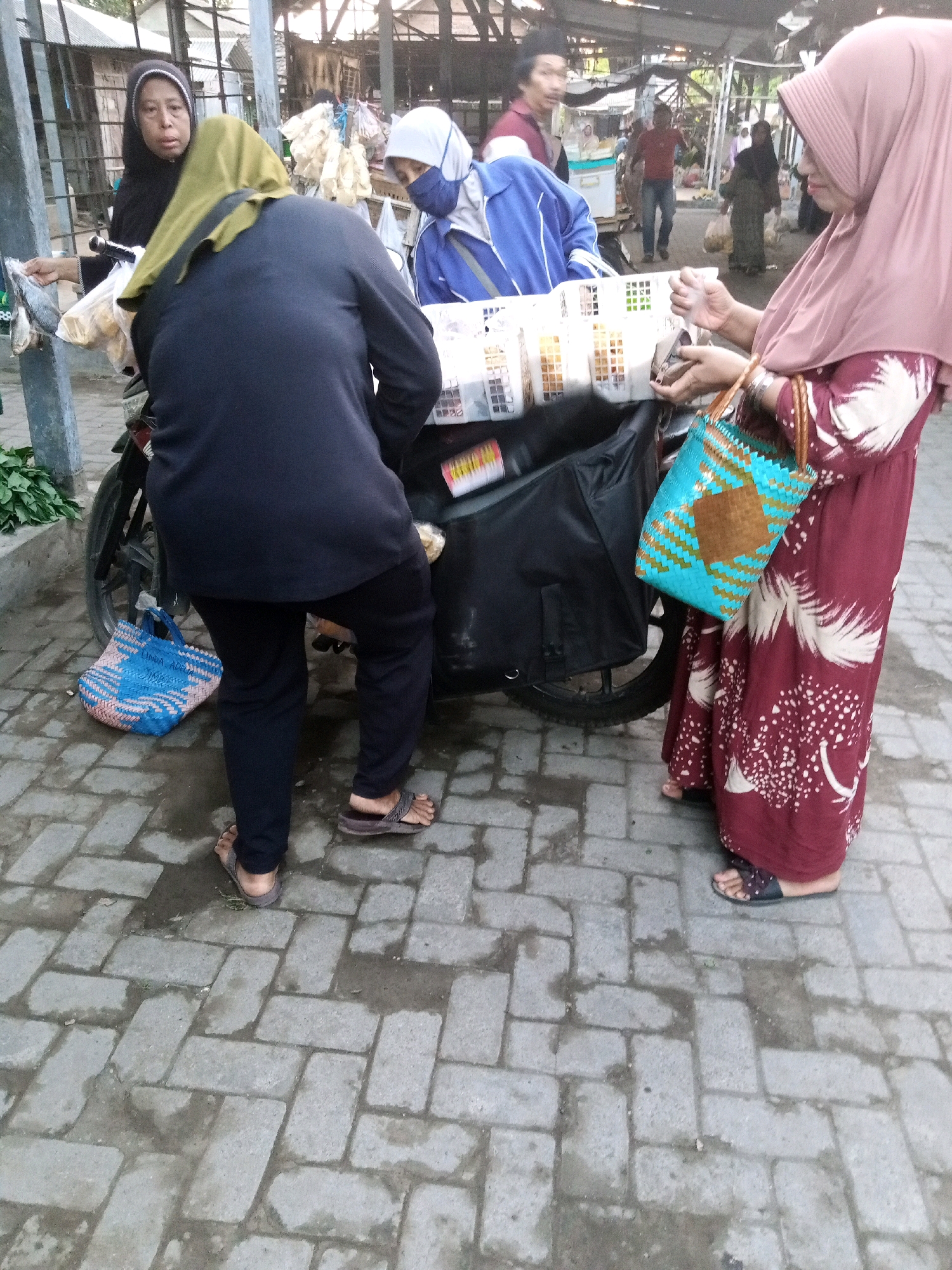 In Ngronggot, Indonesia, on a paved street with a patterned brick surface, several individuals are gathered around a food cart. A woman wearing a black hijab and black clothing stands on the left, holding a plastic bag with what appears to be dried fish and other food items. She is looking at the camera. To her right, a person in a navy blue jacket and a light green headscarf is facing away from the camera, seemingly arranging items in a white basket attached to the cart. In the background, a person in a red shirt and blue jeans is walking away from the cart, and other figures are visible further in the distance. On the right side of the image, a woman in a pink hijab and a maroon dress with a white floral pattern is holding a woven blue and brown bag and interacting with the cart. The food cart itself is covered with a black tarp and displays various packaged food items in white baskets. The environment suggests a market or street vendor setting.
