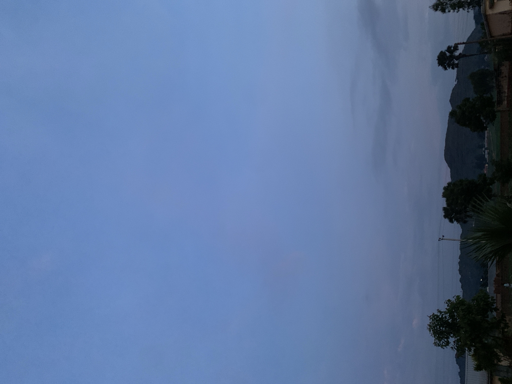 The image depicts a twilight scene over a rural landscape, possibly on the outskirts of Kuru Karama, Nigeria. A vast expanse of pale blue sky dominates the upper portion of the frame, transitioning to muted grey and purple hues near the horizon. Silhouetted against the sky are several trees of varying heights, including what appear to be palm trees on the right side. In the mid-ground, rolling hills form a dark, undulating silhouette. Below the hills, faint outlines of buildings and agricultural fields are discernible. A single utility pole with wires is visible amongst the trees. The overall impression is one of a calm, natural environment at the end of the day.