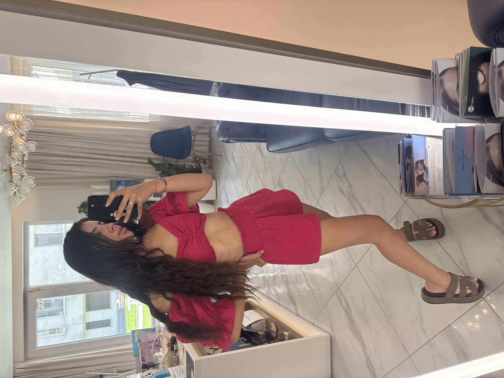 A young woman with long, dark, wavy hair is taking a selfie in a mirror. She is wearing a bright pink off-the-shoulder crop top with ruching detail and matching high-waisted shorts. She has on brown platform sandals. Her left hand is holding a smartphone, and her right hand is on her hip. She is standing in what appears to be a modernly decorated interior space, possibly a salon or boutique. The room has tiled floors, sheer curtains, and a large mirror that reflects the scene. In the background, through a window, a building exterior is visible. To the left of the woman, there is a white display shelf with various products. To the right, a tall mirror with integrated lighting is present. There is a blue chair and a dark sofa visible in the reflection. The overall impression is one of a well-lit and contemporary setting. The image does not contain information that would allow for localization to Plovdiv, Bulgaria.