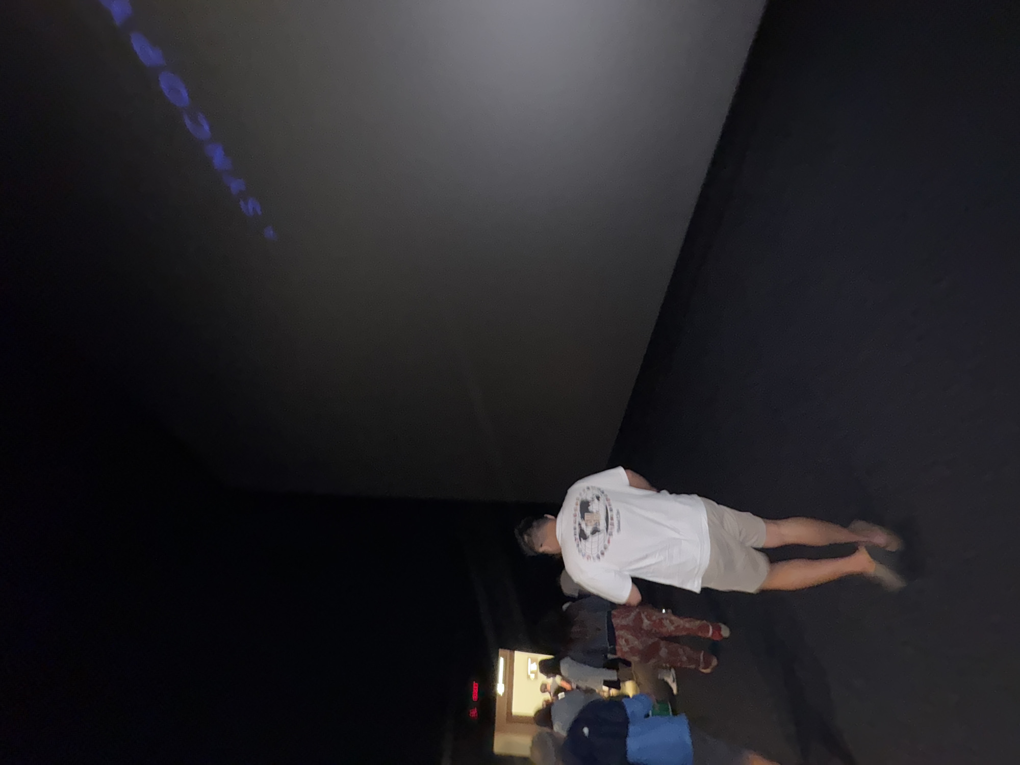 A group of people are walking in a dimly lit corridor toward a large, blank screen in what appears to be a cinema or a presentation hall. In the background, a title or logo with the words "A SYNCOPY" is projected onto the screen in a blue, glowing font. The people are dressed casually, with one man in the foreground wearing a white t-shirt and khaki shorts. The corridor has dark carpeting and the lighting is minimal, creating a sense of anticipation or mystery. The overall scene suggests people are about to enter a screening or presentation. The location in Denpasar, Indonesia is not specified in the image.
