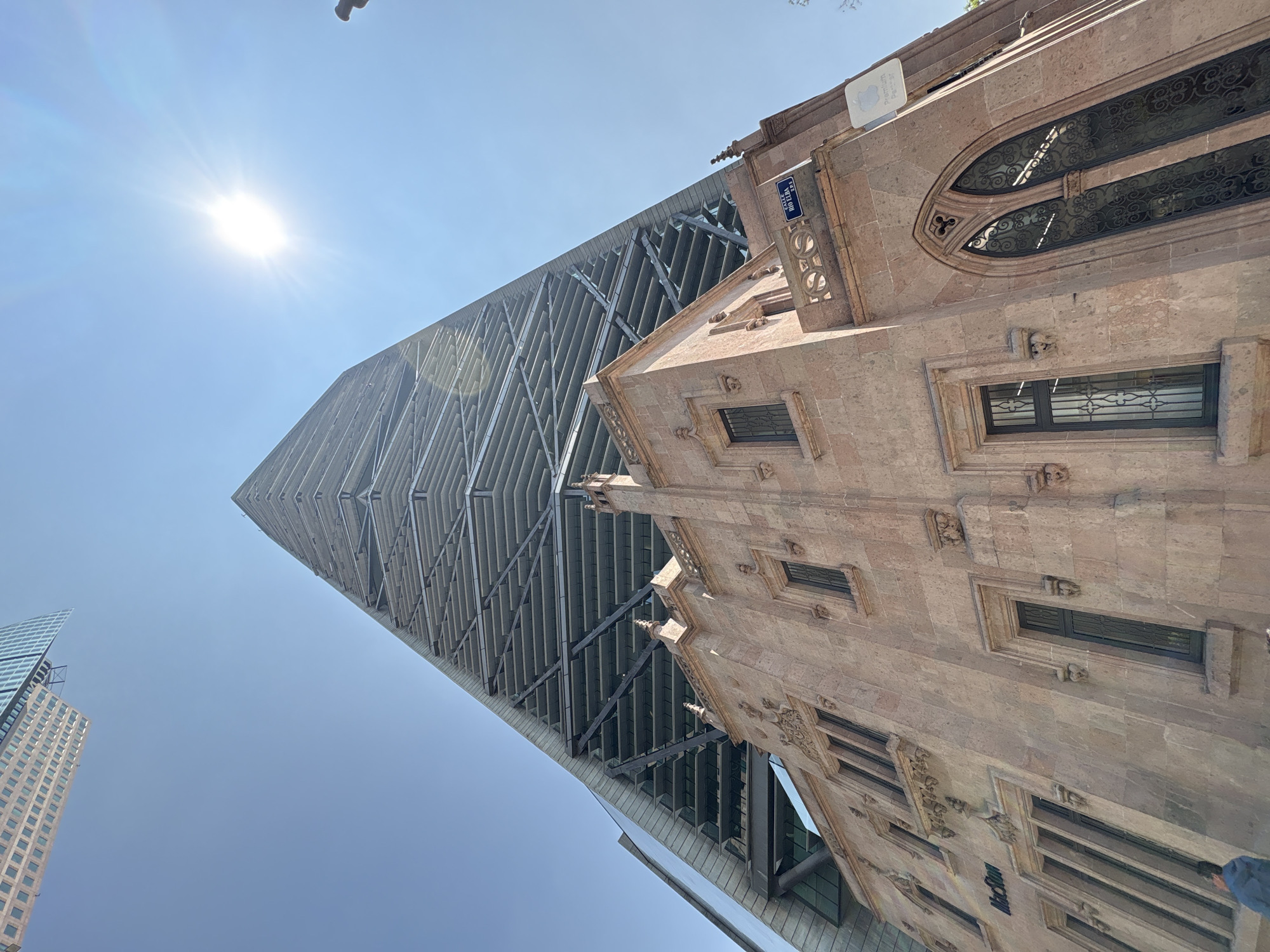 The image, captured with a strong upward perspective, presents a striking contrast between modern and historical architecture in Mexico City under a clear, bright blue sky. A dazzling sun with visible lens flares shines in the upper left, illuminating the scene.
On the left side, a towering, contemporary skyscraper ascends towards the sky, characterized by its intricate, grid-like facade that tapers to a point. Its structure appears modern and sleek. To the right, an older, richly detailed stone building stands, showcasing historical architectural styles. This building features multiple windows adorned with decorative wrought ironwork, pointed arches, and ornate stone carvings that suggest Gothic or Baroque influences. Small, sculptural elements resembling gargoyles are also visible along its cornices.
In the far left background, a glimpse of another modern high-rise with a glass and grid pattern is visible. The pronounced upward angle of the photograph causes the buildings to appear dramatically diagonal, enhancing the sense of scale and height.
Several pieces of text are visible on the old building. A blue street sign with white lettering reads "CALLE" and "VETLAO". Above this, a white sign, possibly associated with an authorized reseller, displays "Premium Partner" followed by "P8-737". Lower down on the stone facade, another sign clearly reads "McCau".