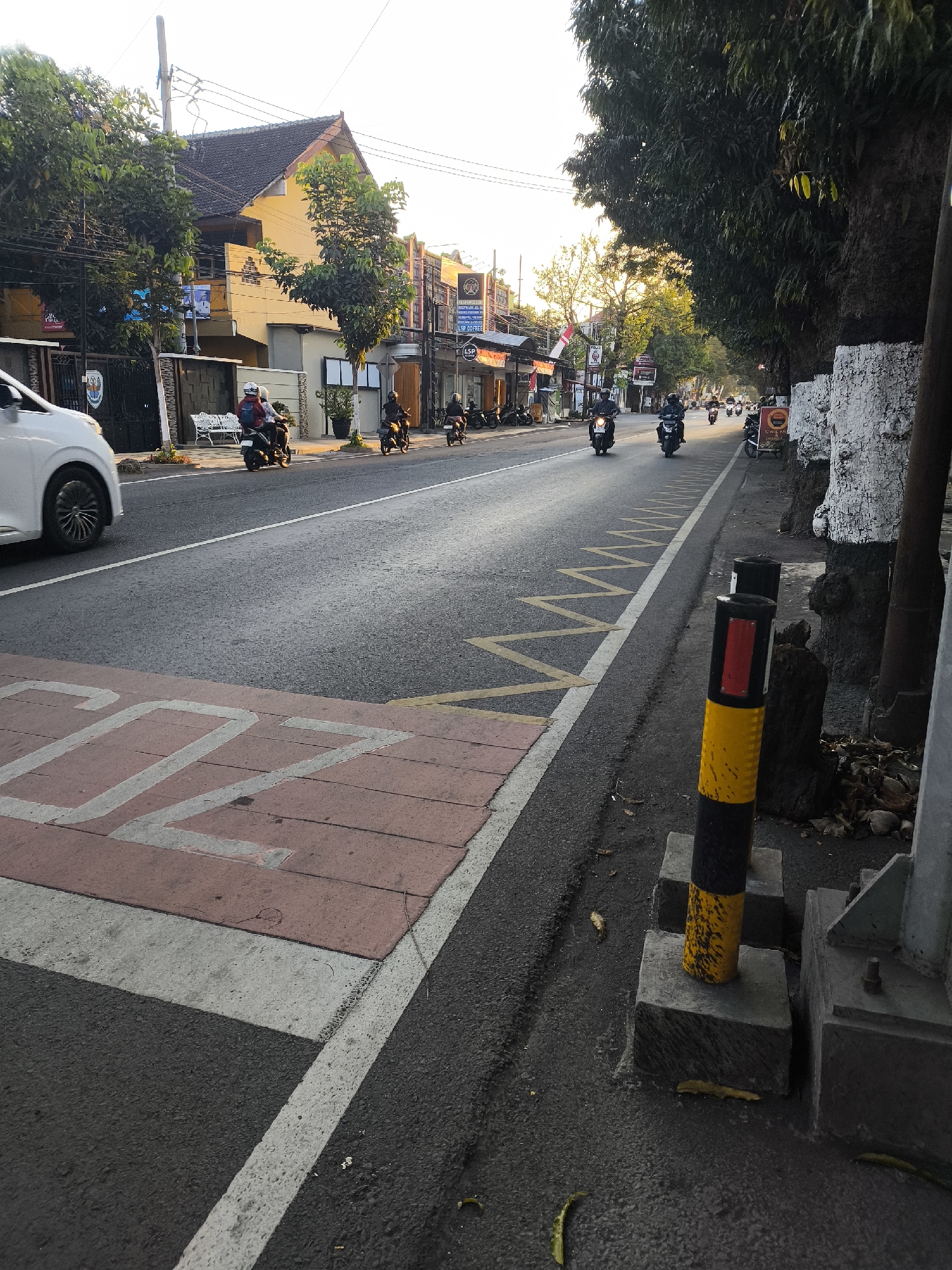 This is a street scene in Citrodiwangsan, Indonesia, with a focus on the road and the pedestrian crossing. The road is paved and appears to be a two-lane street. On the left side of the road, there is a white car and a pedestrian crossing with pink and white markings. Further down the road, several people are riding motorcycles, with some appearing to be in traffic. The buildings on the left side of the street are a mix of styles, with some featuring yellow facades. Trees line the right side of the road, and a traffic bollard with black and yellow stripes stands near the foreground. The sky is clear and bright, indicating daytime.