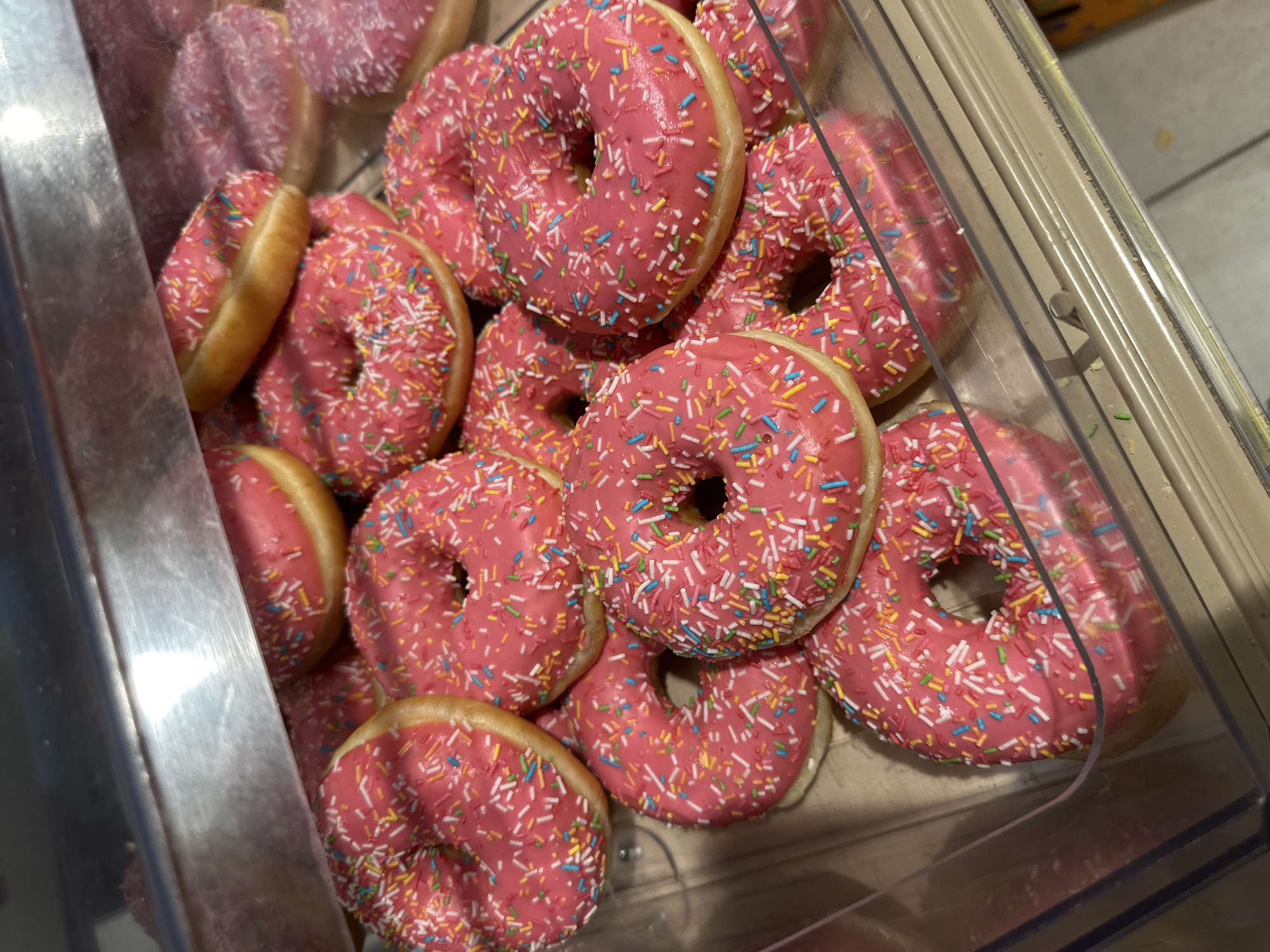 The image displays a close-up of numerous ring-shaped donuts piled within a clear plastic display case. All the donuts feature a bright pink icing and are generously covered with a variety of colorful sprinkles, including white, yellow, blue, green, and pink. The visible parts of the donut dough are a light golden-brown color. The donuts are stacked and overlapping, filling most of the visible container. The setting appears to be indoors, likely in a supermarket, bakery, or a food service area, indicated by the commercial-style display. There are no people visible, and no discernible text can be seen in the frame. The lighting suggests an artificial indoor environment. This scene is located in Omsk, Russia.
