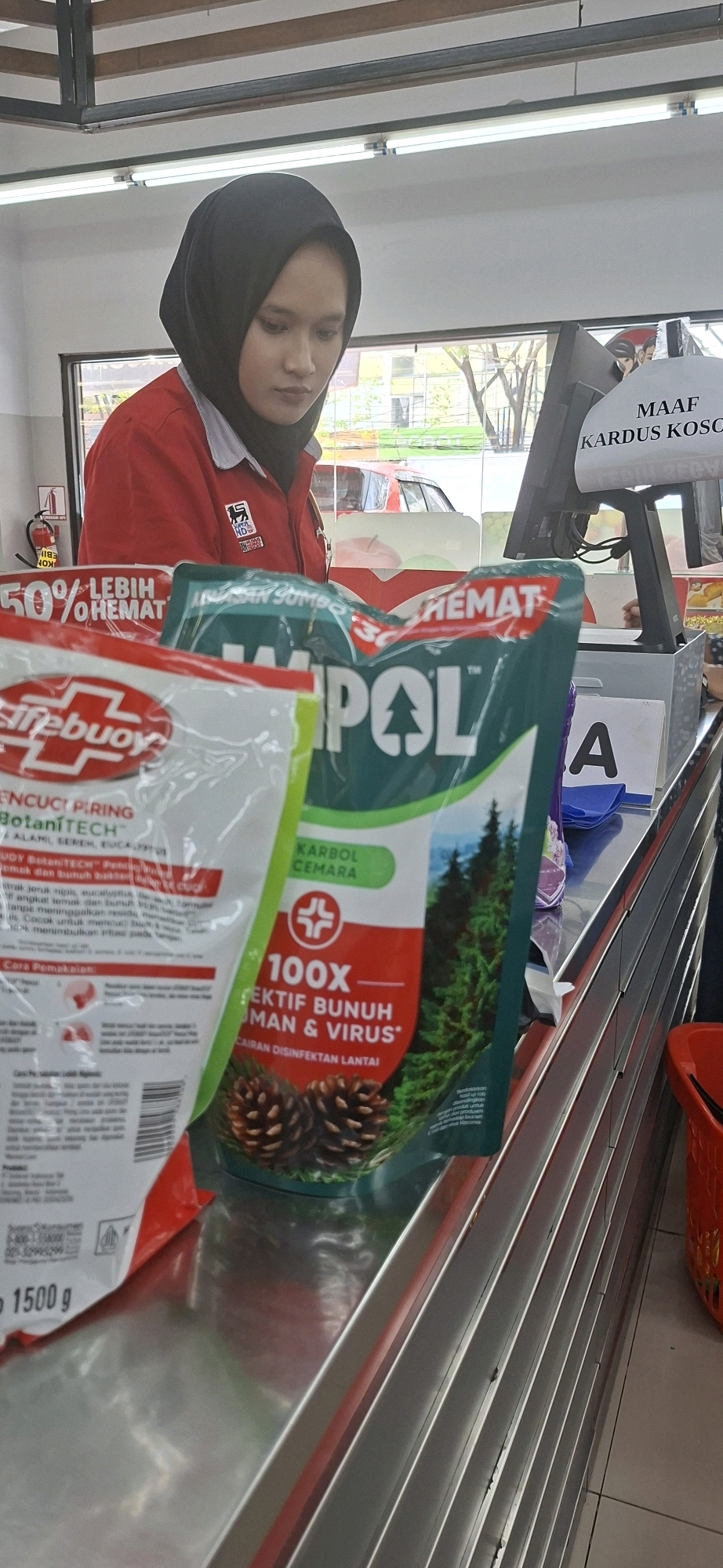 The image captures a scene at a checkout counter, likely in a convenience store or supermarket, in Surabaya, Indonesia. A young woman wearing a black hijab and a red uniform shirt with a logo is visible behind the counter, looking down. She appears to be a cashier or store employee.
In the foreground, several product packages are arranged on the counter. To the left, there is a white and red package for "Lifebuoy" dish soap, with text indicating "BOTANITECH" and "1500 g". To the right of this is a larger, green package of "WIPOL" disinfectant, featuring images of pine trees and pine cones. The text on the WIPOL package includes "KARBOL CEMARA", "100X", and "EFEKTIF BUNUH HUMAN & VIRUS*".
Behind the products, a computer monitor and part of a cash register are visible. A sign on the monitor reads "MAAF KARDUS KOSONG" which translates to "SORRY EMPTY BOX". The environment appears to be indoors, with fluorescent lighting overhead and a window in the background revealing an outdoor scene with trees and a red vehicle. The checkout counter itself is made of reflective metal. The overall impression is a typical retail transaction setting.