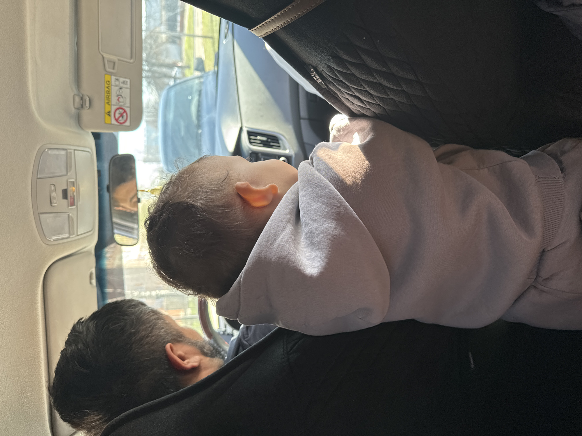 The image captures a candid moment inside a car, likely a stationary or moving vehicle in Chaikovsky, Russia, during daytime under clear weather, as indicated by the bright sunlight streaming through the windows.
The primary subjects are a baby and an adult. The baby, visible from the back of the head and right ear, is dressed in a light-colored, possibly grey, hooded sweatshirt. The baby appears to be asleep or resting, positioned in a car seat or carrier that is mostly out of frame but has a dark, textured back cushion visible. The baby's head is tilted slightly forward, and sunlight illuminates parts of their head and clothing.
An adult, likely male, is partially visible in the foreground, seated in the front passenger seat. Only the back of their head, covered in dark hair with some grey, and a portion of their black clothing are visible. A reflection in the car's rearview mirror shows another adult, presumably the driver, and a glimpse of the outdoor environment, including what looks like a street or open area with distant trees or buildings.
The car's interior is visible, including the cream-colored ceiling, an overhead light console, and a beige sun visor on the passenger side. The sun visor has a prominent yellow warning label with black text that reads "AIRBAG" and features several warning pictograms, including symbols for a child in a front airbag zone, a rear-facing child seat, and a seatbelt. Faintly, "SRS AIRBAG" text is also visible on the side of the car's seat or headrest material. Car vents are also seen in the background. The overall scene suggests a quiet, peaceful journey or pause.